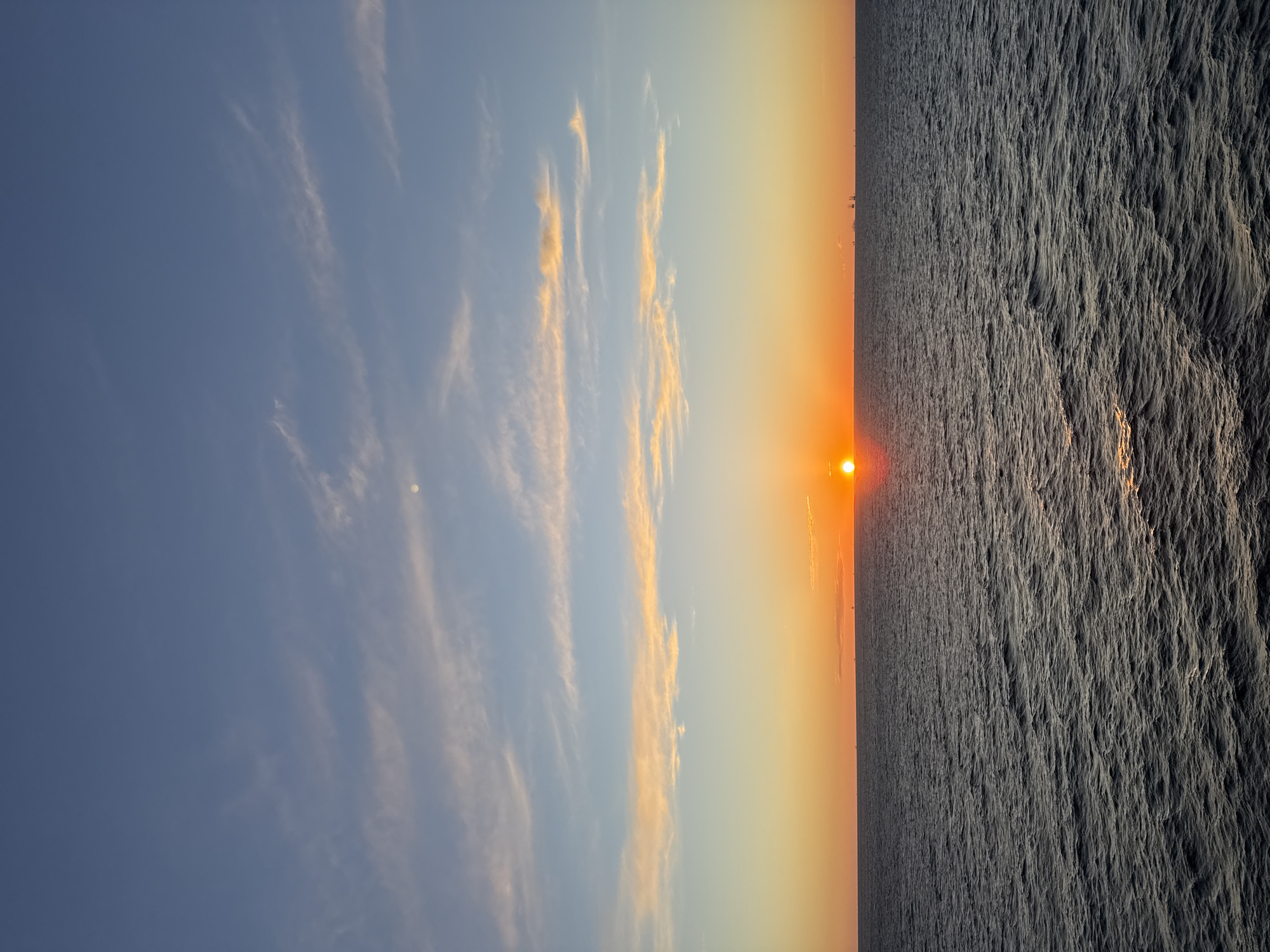 The image captures a sunset over the ocean. The sun is visible on the horizon, appearing as a bright orange orb. The sky transitions from blue with scattered clouds to a yellow-orange gradient near the horizon. The ocean surface appears textured with patterns of light and shadow. The location is Unknown.