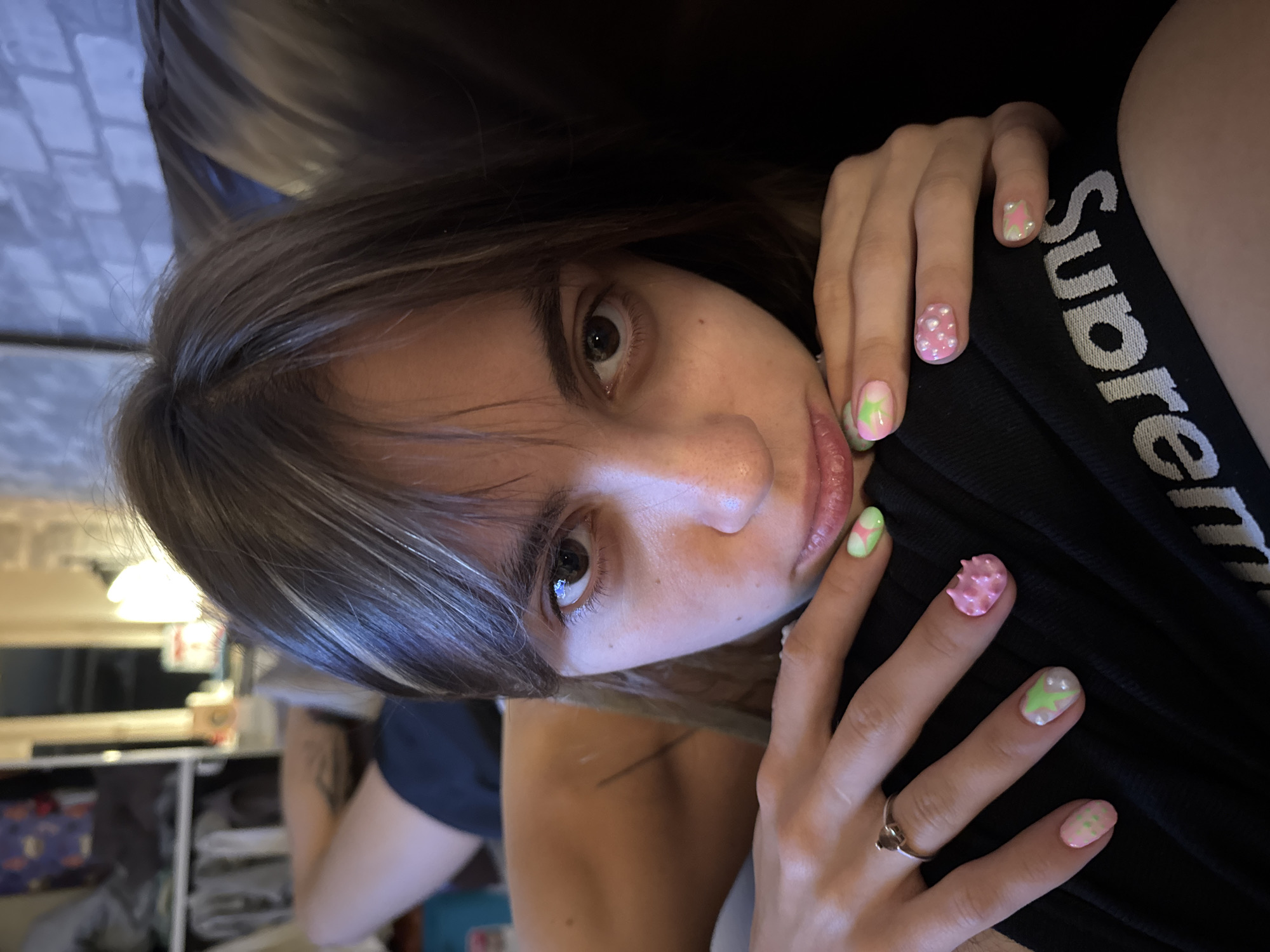 The image features a close-up, rotated portrait of a young person lying down, with their face partially visible. Their eyes are open and looking directly at the camera. They have dark hair that falls across their face. Their hands are in the foreground, with intricately designed fingernails. These nails are decorated with pink and green patterns, including what appear to be flowers and possibly stars, with some nails having a textured, bubbly finish.
The person is wearing a black garment with white lettering that reads "Supreme". A silver ring is visible on one of their fingers. The background is dark and out of focus, with hints of indoor lighting and what looks like furniture or shelving. The lighting appears to be artificial, suggesting the photo was taken indoors, possibly in the evening. The overall mood is intimate and personal, with a focus on the subject's face and hands. There are no overt clues about the weather or specific time of day beyond it being likely evening or night. The location context of Moscow, Russia, is provided externally and is not discernible from the image itself.