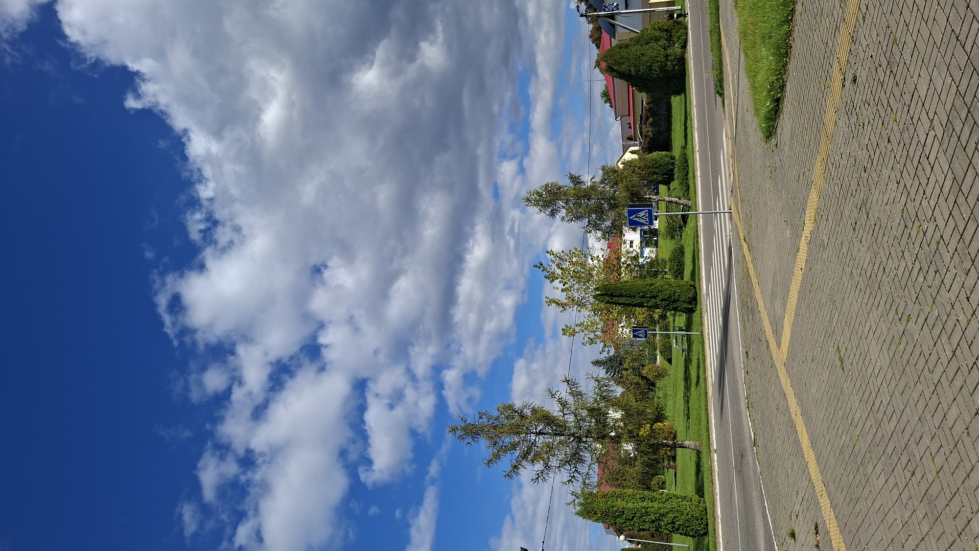The image depicts a paved walkway with tactile paving leading towards a street crossing marked with a pedestrian crossing signal and a white crosswalk. The street runs parallel to a green lawn bordered by trees and buildings in the background. The sky is partly cloudy with a mix of blue sky and gray clouds. The scene appears to be located in an urban or suburban area, possibly in the city of Myadzyel, Belarus, given the context. No people or animals are visible in the immediate foreground. The overall impression is one of a calm, ordinary day in a public space.