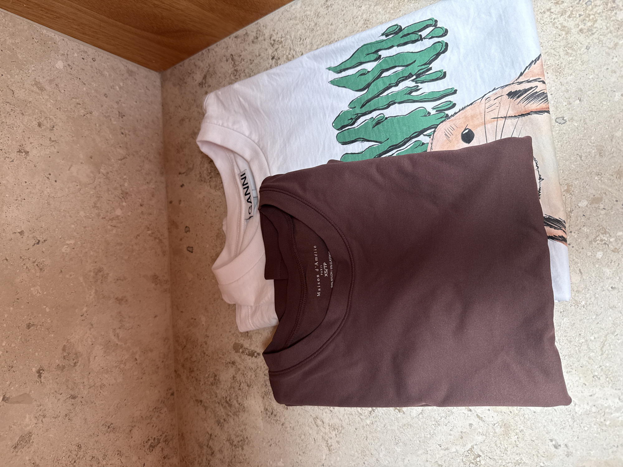 The image displays an overhead view of two folded t-shirts resting on a light-colored, mottled stone-tiled surface. A warm-toned wooden surface forms a corner with the tiled wall in the upper left, suggesting an indoor setting like a shelf or closet.
The top shirt is white and features a graphic design. This design includes abstract green shapes, possibly depicting foliage or a stylized tree, along with a light brown animal-like figure that has a dark eye and a bushy tail, resembling a squirrel or similar creature. Part of the white shirt's brand tag is visible, reading "GANNI".
Beneath the white shirt is a plain, solid-colored t-shirt in a deep brown or maroon hue. Its inner collar tag is clearly visible, identifying the brand as "Maison d'Amélie". The tag also indicates the size as "XS/TP" and specifies the fabric composition as "94% NYLON / 6% ELASTANE".
No people are visible, and no specific activity is taking place; the shirts are simply arranged on the surface. The lighting is even, consistent with an indoor environment. The textured tiles and wooden element create a natural, earthy backdrop for the clothing items.