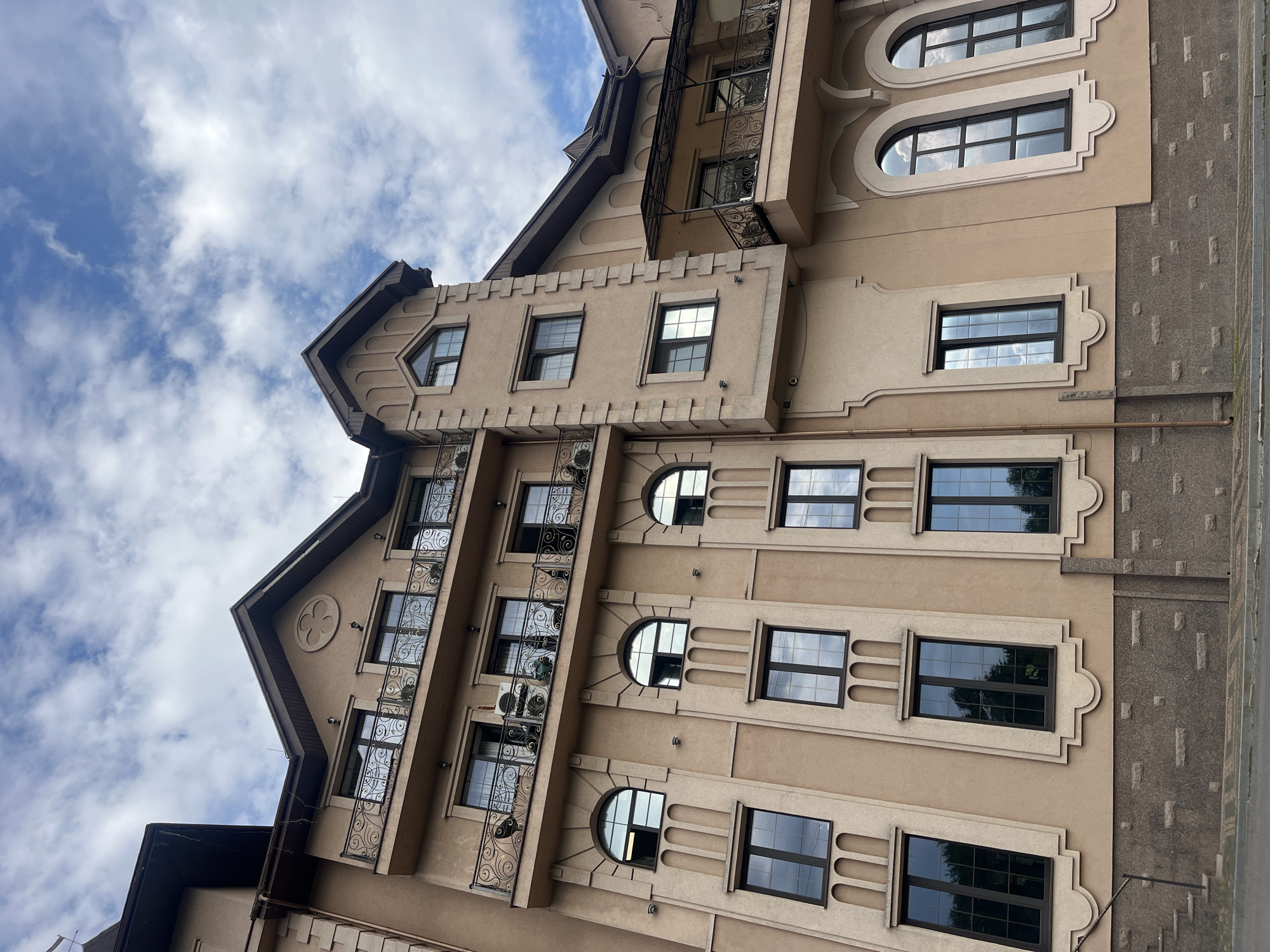 The image captures a detailed, tilted view of a multi-story building against a partly cloudy sky. The architecture is elaborate, featuring a light brown or beige facade with dark brown accents on the roofs, window frames, and decorative elements.
The building incorporates various window designs, including rectangular, semi-circular, and arched shapes, many adorned with decorative surrounds and cornices. Multiple balconies with intricate, ornate black metal railings are visible, adding to the building's sophisticated aesthetic. One prominent gable on the upper left features a circular, quatrefoil-like decorative motif. Sections of the exterior wall, particularly on the right side, are finished with a contrasting grey, textured stone-like material.
An air conditioning unit can be seen mounted below one of the balconies. Drainage pipes run down the building's exterior. The sky is blue with scattered white clouds, indicating daytime and fair weather. The overall style suggests an eclectic or possibly neo-Gothic architectural influence. No human activity or visible text is present in the image. The context for the location is Kamianytsia, Ukraine.