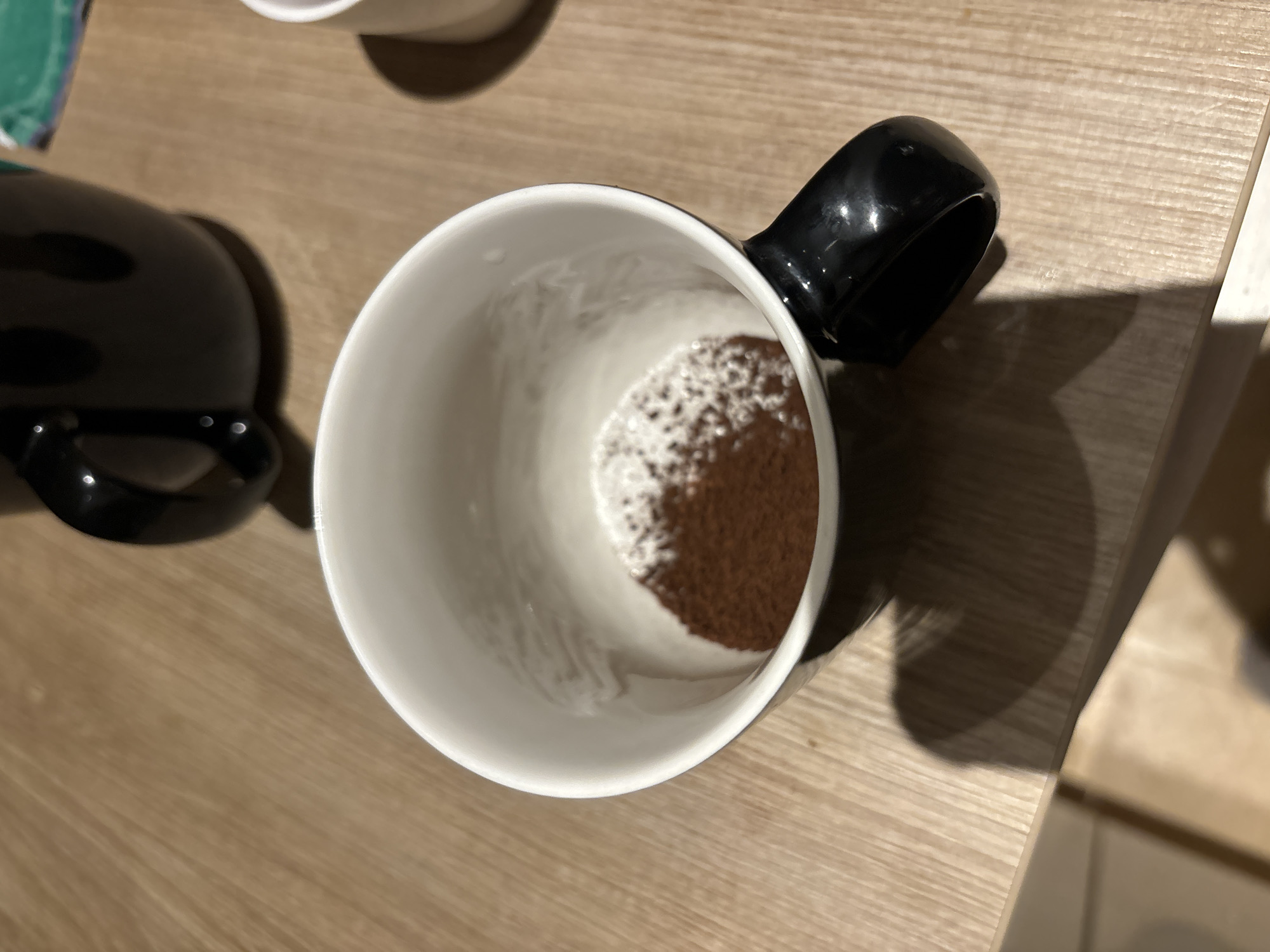 The image shows a white mug with a black handle sitting on a light brown wooden surface. Inside the mug, there is a brownish powder residue. To the left of the mug, part of another black object, possibly another mug or container, is visible. The wooden surface appears to be part of a table. The photograph seems to be taken from above, looking down into the mug. The specific location within Budva, Montenegro, cannot be determined from the image alone.
