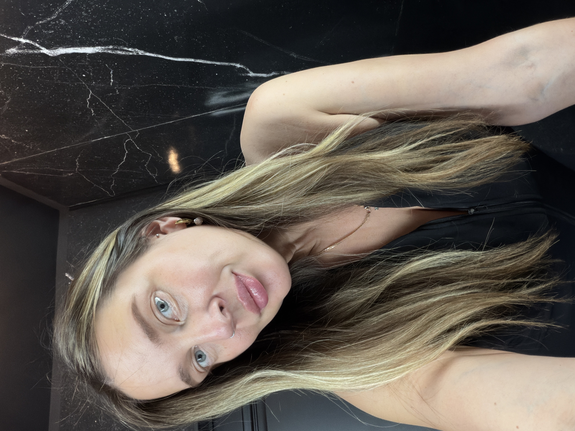A woman is pictured in a selfie, her head tilted towards the camera. She has long, blonde hair with subtle highlights, and her eyes are a striking blue. She is wearing a black top and a delicate gold necklace. A small hoop nose ring is visible, as are earrings. The background appears to be a dark, reflective surface, possibly marble, with a prominent white vein running through it. The lighting suggests an indoor setting, likely a bathroom or a similarly well-lit area, with no indication of time of day or weather. The woman's expression is neutral, with slightly parted lips. The overall tone is casual and intimate, characteristic of a personal selfie.