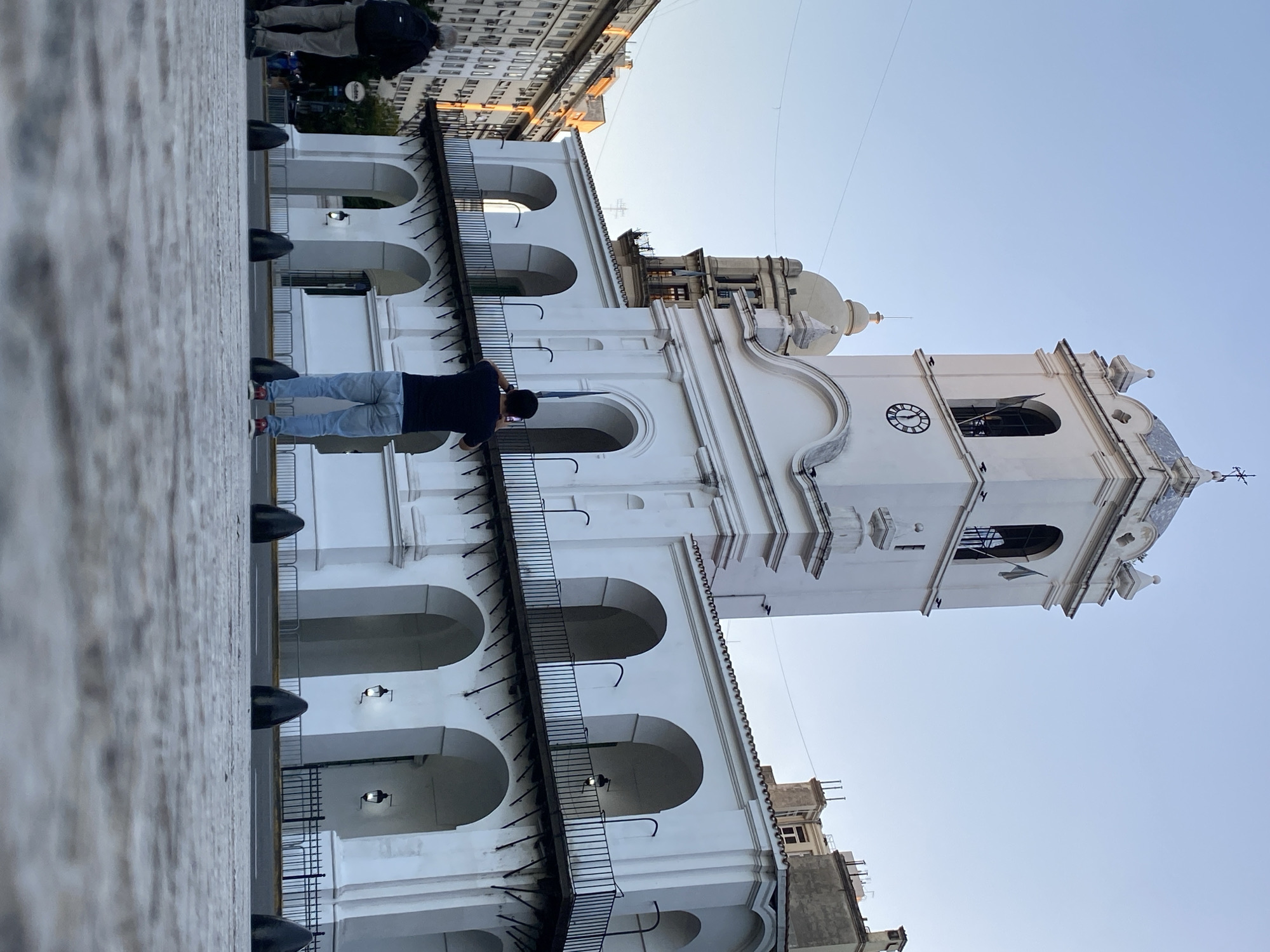 A low-angle, portrait-oriented photograph depicts the Cabildo de Buenos Aires, a historic building in Buenos Aires, Argentina, against a pale blue sky. The Cabildo, rendered in white, features a two-story arcade with arched openings on both levels. Black iron railings line the balconies. A clock tower rises from the building's corner, displaying a clock face. A cross is affixed to the top of the tower. A person wearing a dark shirt and blue jeans stands on the stone sidewalk in front of the Cabildo, looking up at the building. Another person stands near the sidewalk's edge. Modern buildings are visible in the background, suggesting a juxtaposition of old and new architecture. Black bollards line the sidewalk, presumably to prevent vehicles from driving onto the pedestrian area.