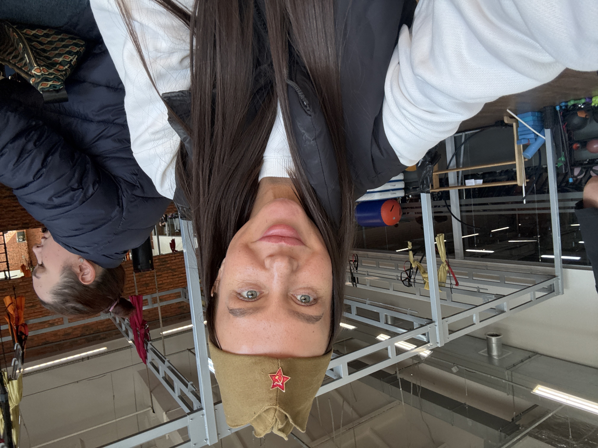 A selfie depicts a woman wearing a Soviet Army garrison cap with a red star, against a backdrop of an indoor facility with a metal structure overhead. To the left of the woman, another individual with tied-back hair, wearing a puffy black jacket, is partially visible. The indoor facility has metal framework suggesting a gym or training area. The image was presumably taken indoors in Chaikovsky, Russia.