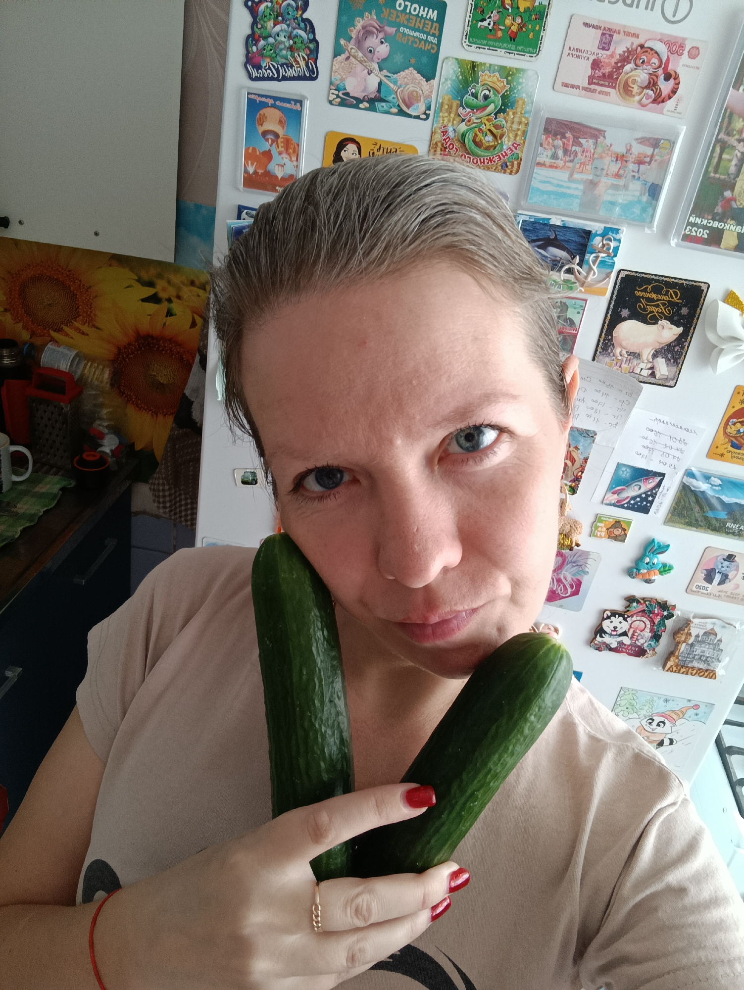 A woman with short gray hair, wearing a light beige t-shirt, holds two green cucumbers near her face. She has blue eyes and red nail polish. She is standing in a kitchen, with a refrigerator behind her covered in magnets and notes. A sunflower-patterned surface is visible to the left of the refrigerator.
The presence of a magnet of the city suggests this photo was taken in Chaikovsky, Russia.
