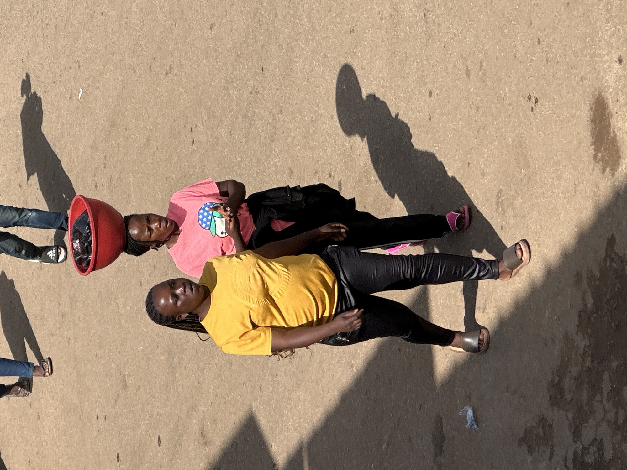 Two women are walking on a dirt road in Bukuru, Nigeria, under bright sunlight. The woman in the foreground, on the left, wears a yellow t-shirt and black leggings, with her hair in braids. She is walking with her left foot slightly forward. The woman behind her, on the right, wears a pink t-shirt and black leggings, and is carrying a red bowl filled with black items on her head. Her right foot is slightly forward. Shadows are cast on the road, indicating a sunny day. The road surface is a light brown color with small pebbles and some wet patches. There is a crumpled piece of white paper on the road to the left of the foreground woman. In the background, blurred legs of other individuals are visible, also walking.