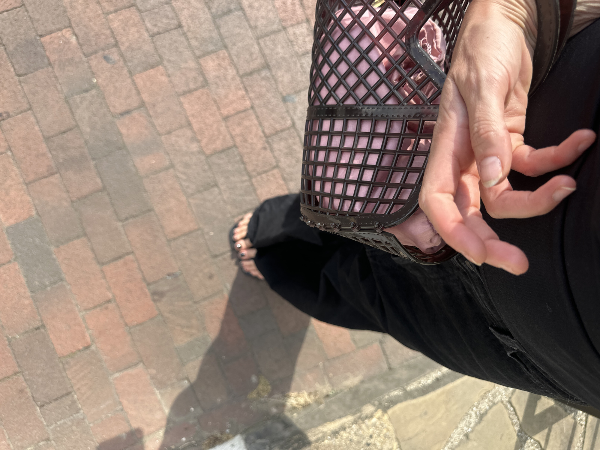 The image shows a top-down view of a person's lower body and hands, holding a pink woven handbag. The person is wearing black pants and sandals with dark nail polish. They are standing on a brick pathway, with a shadow cast to their left. The pathway appears to be in an outdoor urban setting. Without further information, the specific location in Tunbridge Wells, United Kingdom cannot be determined.