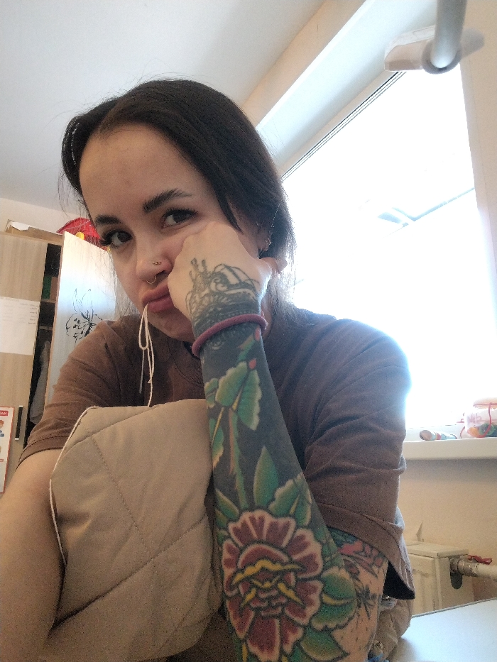 The image displays an individual, identified as a young adult female, situated indoors. The location is stated as Noviy, Russia; however, this specific geographic detail cannot be corroborated by visual elements within the photograph. The subject has dark hair, dark eyes, and a fair complexion. Facial piercings include a silver septum ring and a silver stud on the left nostril. The lips are pursed downwards, with a light-colored string or thread emerging from the mouth. The left arm, visible in the foreground, features a full sleeve tattoo characterized by a dark background adorned with green leaves and prominent red and yellow flowers, including a large floral design on the lower forearm. A red elastic band encircles the left wrist, and the back of the left hand displays a tattoo resembling an ornate crown. The left hand is positioned against the side of the face. The individual wears a brown long-sleeved shirt.
The background on the right is dominated by a large window emitting strong, overexposed backlighting, obscuring external details. A white window frame and a light-colored latch mechanism are discernable. Below the window, a white windowsill holds several small, colorful objects contained within a clear vessel, alongside a white radiator or heating pipe component. To the left of the subject, a light-colored wooden cabinet or shelf unit is partially visible, with a piece of paper or material featuring a floral outline attached to its surface. The ceiling above is light-toned.