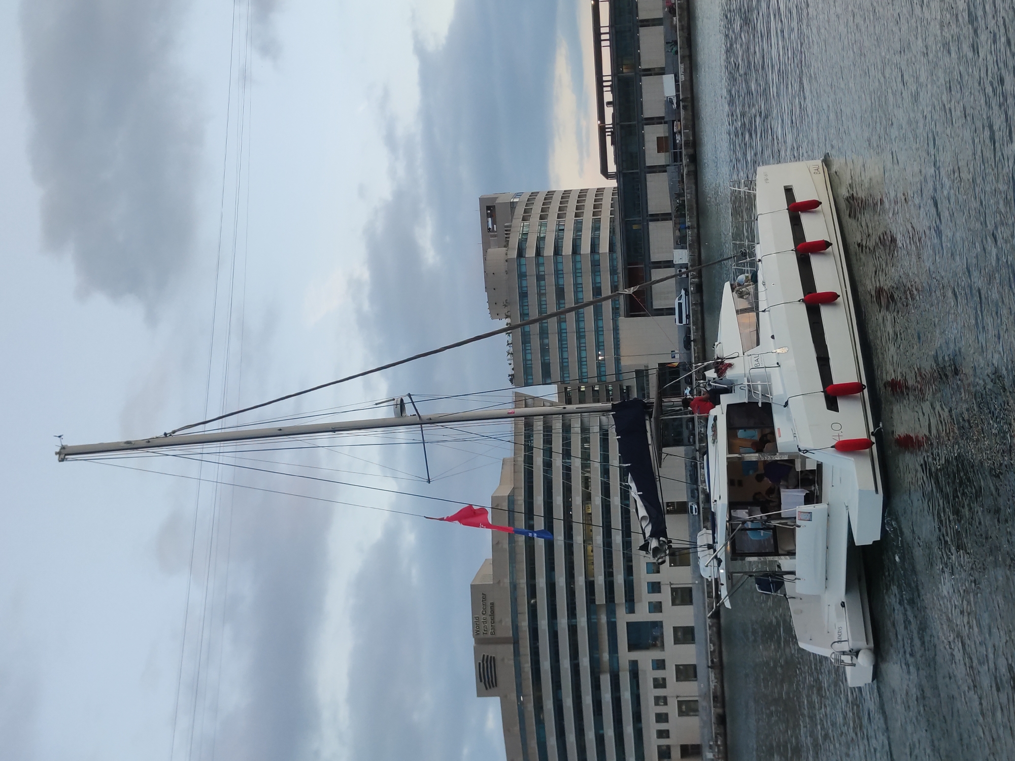 The image depicts a white catamaran sailing on the water with a cityscape in the background. The buildings in the background appear to be part of a port or harbor area. One prominent building in the background has "World Trade Center Barcelona" written on it, indicating the location as Barcelona, Spain. The catamaran has a mast and sails, and several people are visible on its deck. Red fenders are attached to the sides of the boat. The sky is overcast with clouds. The water is calm with ripples.