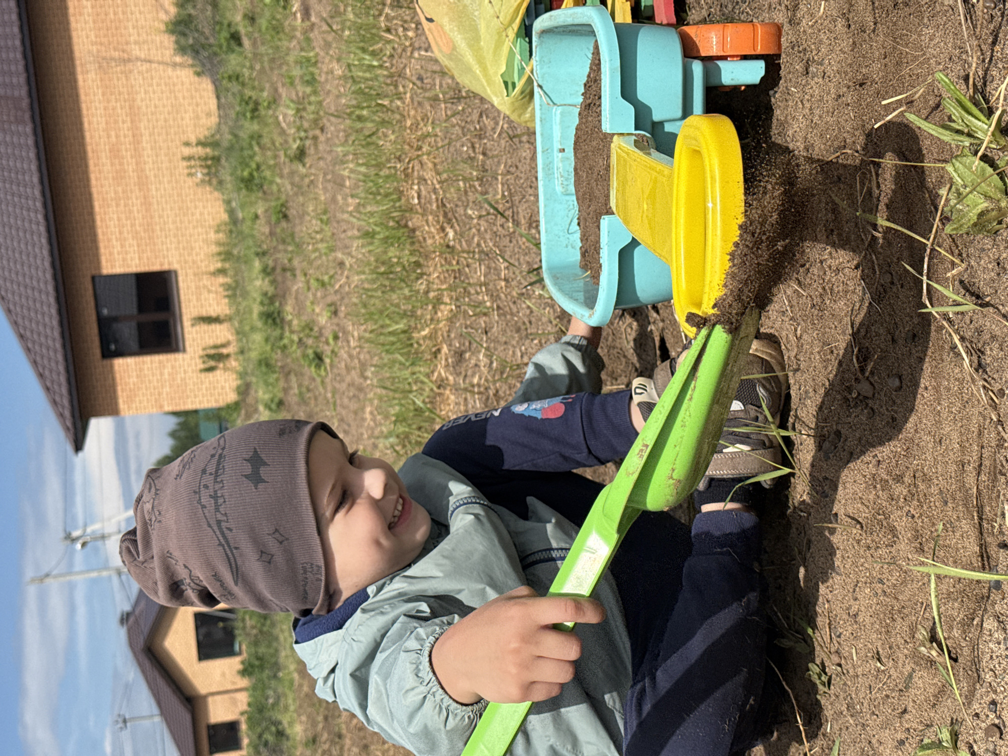A young child, presumably male, wearing a gray patterned beanie, light green jacket, and dark blue pants, sits on the ground outdoors and plays with toys. He holds a green shovel in his hands and looks to his left with a smile. In front of him, a blue and yellow plastic toy, possibly a dump truck or construction vehicle, sits on the ground, filled with dirt or sand. The ground is predominantly brown dirt with scattered patches of green grass. In the background, a brick house with a dark window and a brown roof, along with another house further away, can be seen. The sky is partly cloudy, suggesting a bright day. The location is in the city of Chaikovsky, Russia.