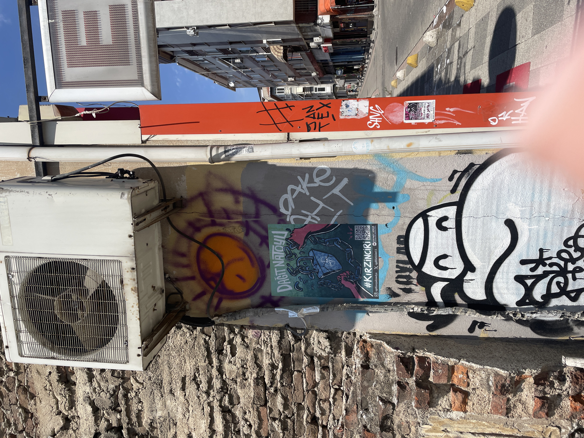 An outdoor, eye-level, medium shot captures a close-up view of graffiti and urban detritus on a weathered brick wall and a white vertical pipe on a sunny day in Kadıköy, Turkey. An outdoor air conditioning unit is mounted above the graffiti. The wall is covered with various spray-painted tags and images, including a stylized sun with a face, an abstract design with purple and orange colors, and a poster with the text "#KIRZINCI'RI" and a QR code. A red pole with more graffiti and stickers stands to the right of the pipe. In the background, a street with buildings and parked cars is visible. The scene is filled with the vibrant colors and textures of urban street art and decay.