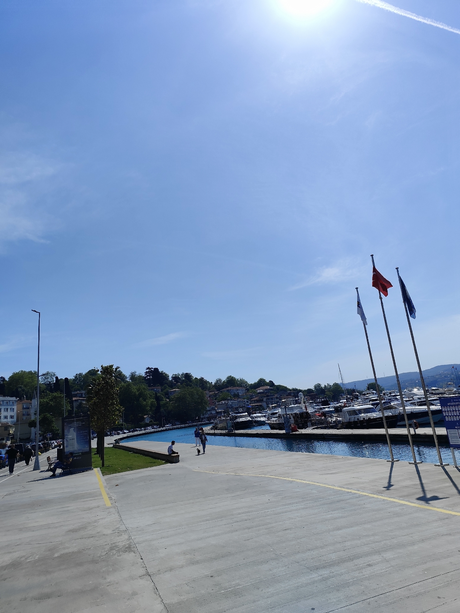 The image captures a sunny day along the waterfront in Sarıyer, Turkey. The scene features a concrete promenade with a yellow painted stripe near the edge, where three flagpoles stand, displaying flags of red, white, and blue colors. In the background, a marina is filled with boats and yachts. The water is reflecting the clear blue sky. A tree-covered hillside forms the backdrop of the marina. People are seen walking and sitting along the waterfront. On the left of the image, a street light pole stands near a bus stop. A man is sitting on a bench next to an advertisement display. The sky is a clear blue with the sun shining brightly. A faint contrail is visible near the top right corner.