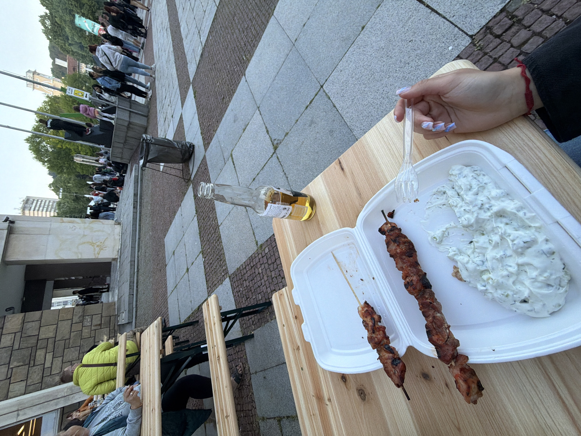 The image shows a person holding a fork over a styrofoam container of food consisting of meat skewers and a white sauce, possibly tzatziki, on a wooden table. There's an empty beer bottle also on the table. The table is outdoors with a view of a brick and gray stone pavement area where a line of people is visible. In the background there are trees and what appears to be a building. The scene appears to be set in a public space with casual dining arrangements. From an approximate geolocalisation, the location could be Gabrovo, Bulgaria.