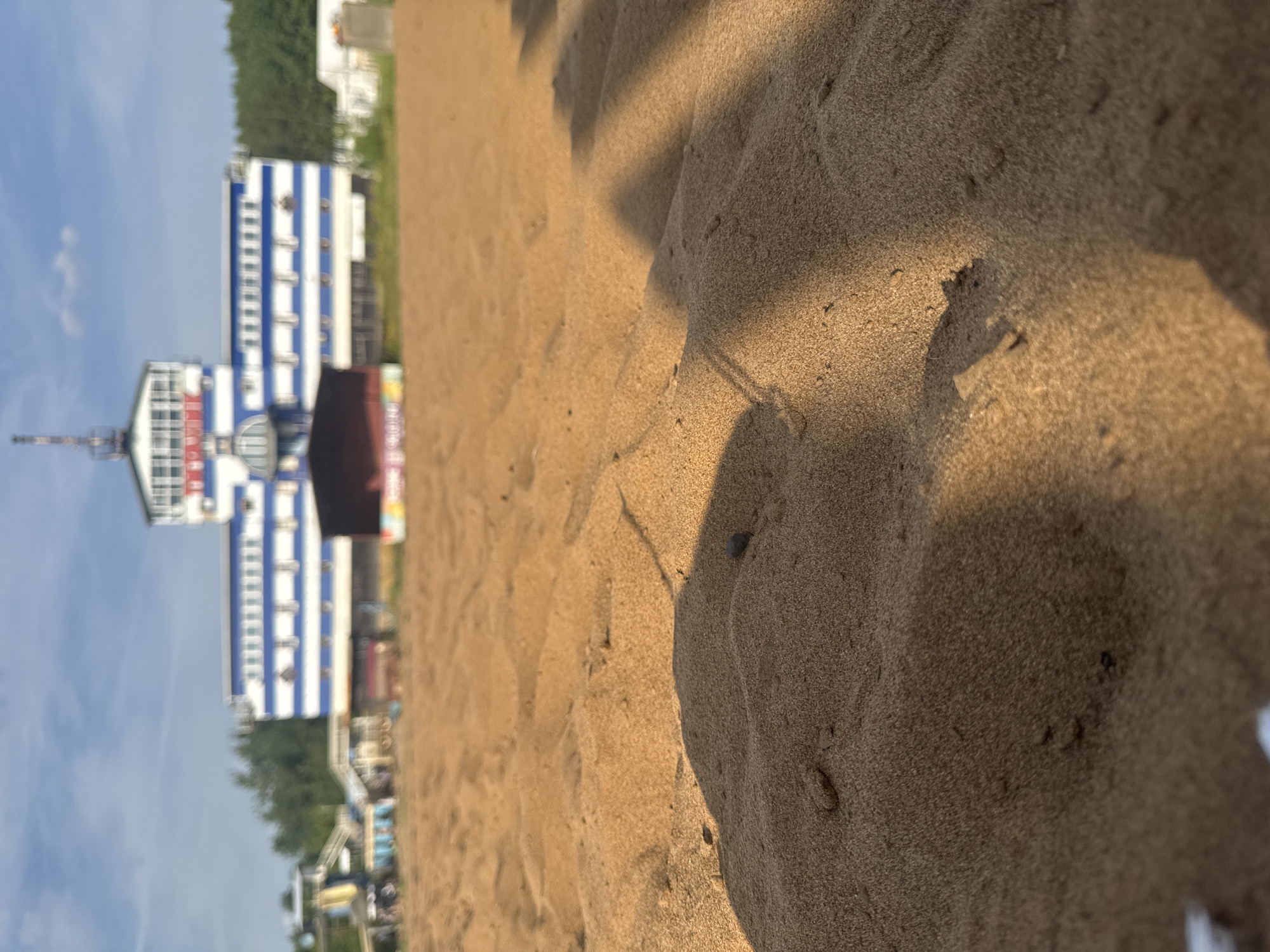 This media file captures a close-up, low-angle perspective of a sandy beach in Kirov, Russia. The foreground is dominated by the texture of the sand, showing ripples and small pebbles, with a shadow cast across it, suggesting it is a sunny day. In the background, a white and blue striped building with multiple stories is visible, appearing to be a hotel or a resort. A tall antenna-like structure is attached to the left side of this building. Further into the background, there are hints of trees and other smaller structures, potentially part of a beachside establishment or park. The sky is blue with a few wispy clouds. The scene suggests a daytime setting, likely during pleasant weather suitable for a beach visit. No specific activity or emotion is discernible due to the focus on the sand and the distant, blurred buildings. No visible text is present in the image.