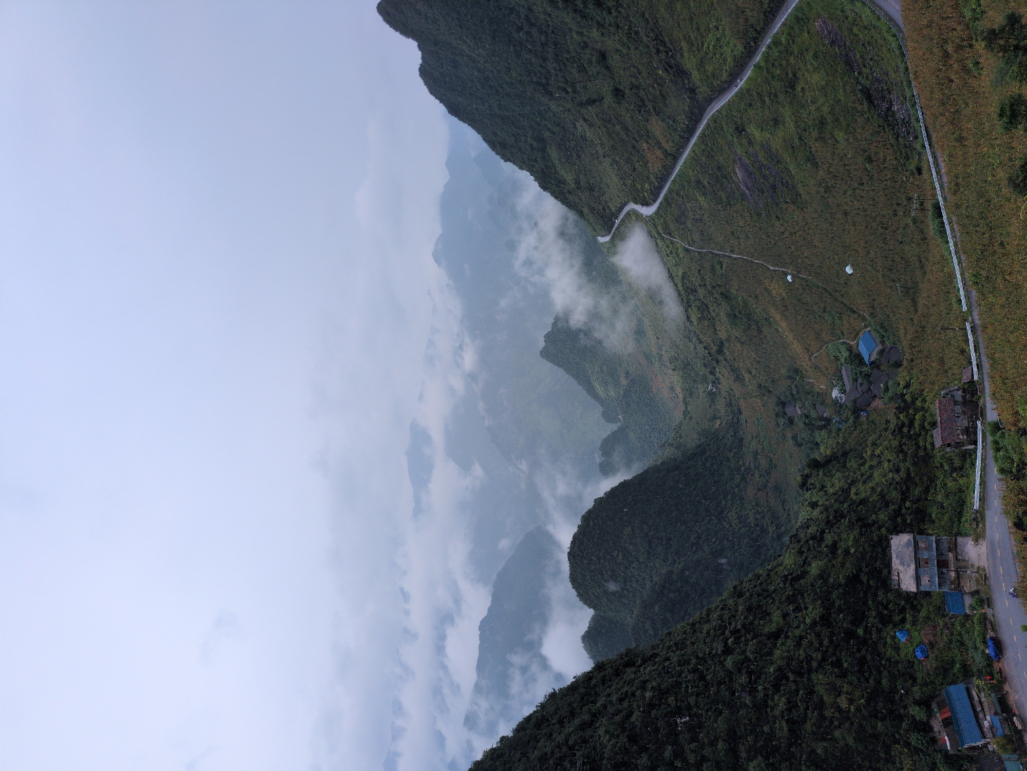 An aerial, high-angle, wide shot captures a misty, mountainous landscape in Đồng Văn Commune, Vietnam, under an overcast sky. The image is dominated by layers of verdant, steep mountains shrouded in fog, creating a sense of depth and mystery. A winding asphalt road snakes its way up and across the mountainside, with a visible car traveling on it. In the valley, a small cluster of houses with blue and red roofs are nestled among the lush vegetation. Several blue tents are also visible near the houses. The terrain is densely covered with trees and vegetation, with some areas appearing to be cultivated fields. The overall atmosphere is serene and remote, highlighting the natural beauty of the region.
