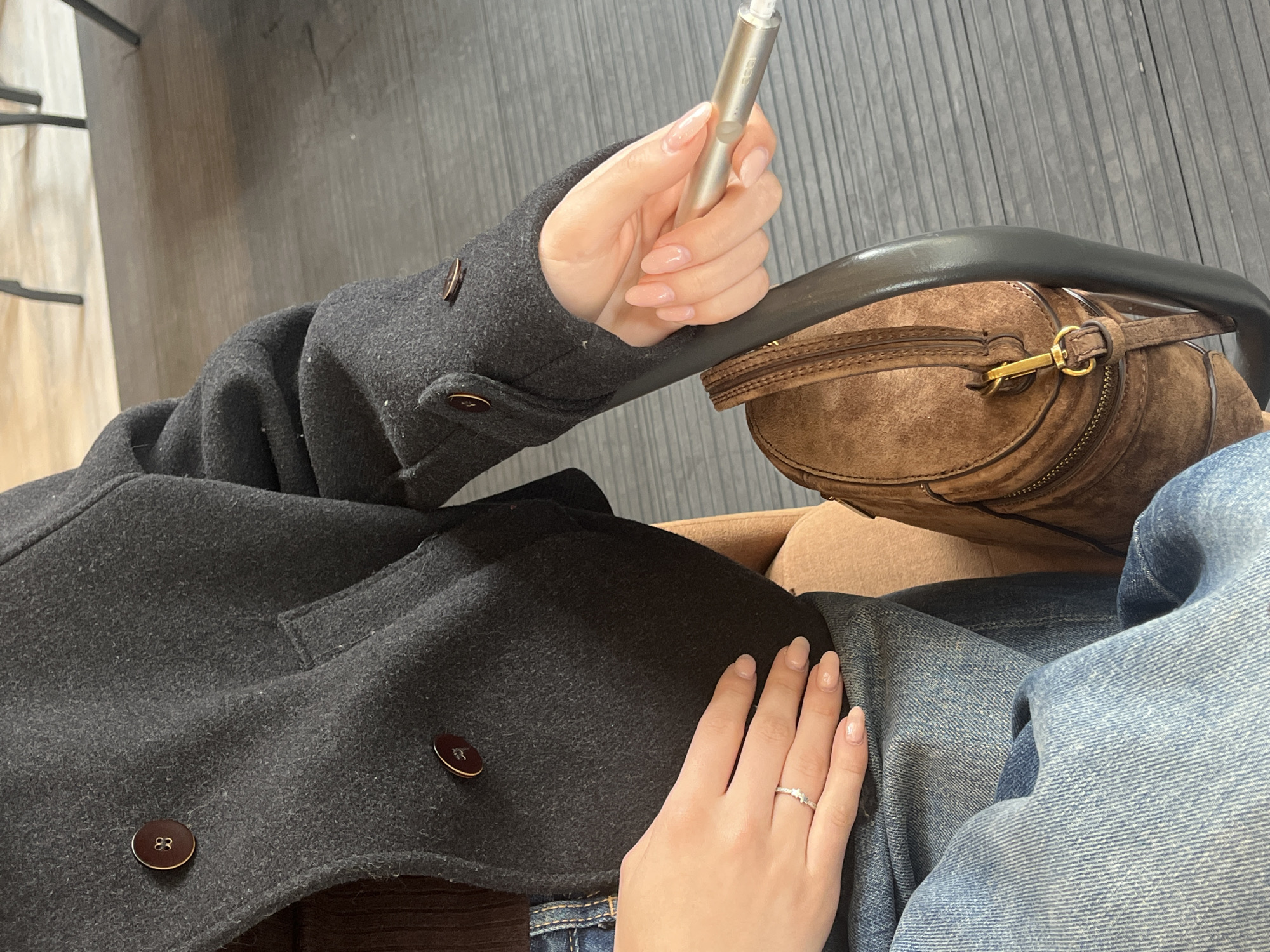 The image provides an overhead view of a person seated indoors in Plovdiv, Bulgaria, focusing on their torso, hands, and immediate belongings. The individual is wearing a dark gray wool-blend coat featuring prominent brown buttons, some faintly marked with the number "83." Beneath the coat, blue denim jeans are visible.
The person's hands are neatly manicured with long, light pink/nude polished nails. The right hand holds a slender, silver-colored device, visibly branded "IQOS," indicating a heated tobacco product. The left hand rests casually on the lap, adorned with a delicate silver-colored ring featuring a small, clear stone on the ring finger.
A brown, suede-like shoulder bag or pouch with gold-colored hardware, including a zipper and a clasp, is situated next to the person's right side, partially obscured by their arm and coat. The seating appears to be a light brown upholstered chair or bench. In the background, there is dark gray or black paneling with a distinct ribbed texture, suggesting a modern interior setting like a cafe, lounge, or waiting area. The overall scene conveys a sense of calm and leisure.