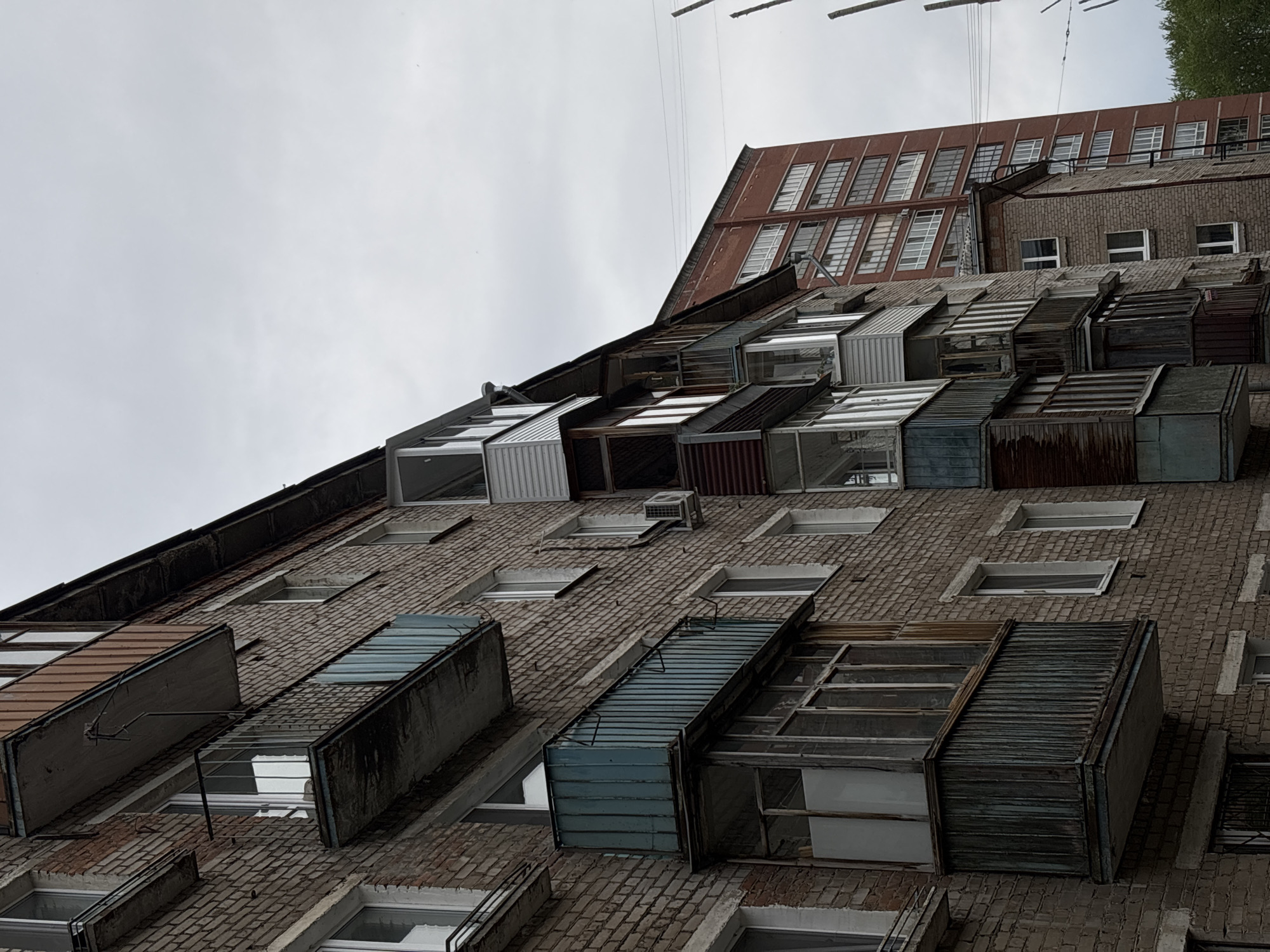 The image depicts the exterior of a building in Perm, Russia, notable for the balconies or extensions attached to the facade. The building's facade is made of brick and the balconies jut out in a non-uniform manner, constructed from a mixture of materials including what appears to be metal siding, corrugated materials, and window frames. Some of the balconies enclose extensions from the windows, while others appear to be open-air spaces. The building is angled upwards in the frame, offering a low-angle view against a gray, overcast sky. A taller, red-brick building is visible in the background, featuring multiple rows of windows. Overhead, wires are strung above the buildings, adding to the urban setting.
