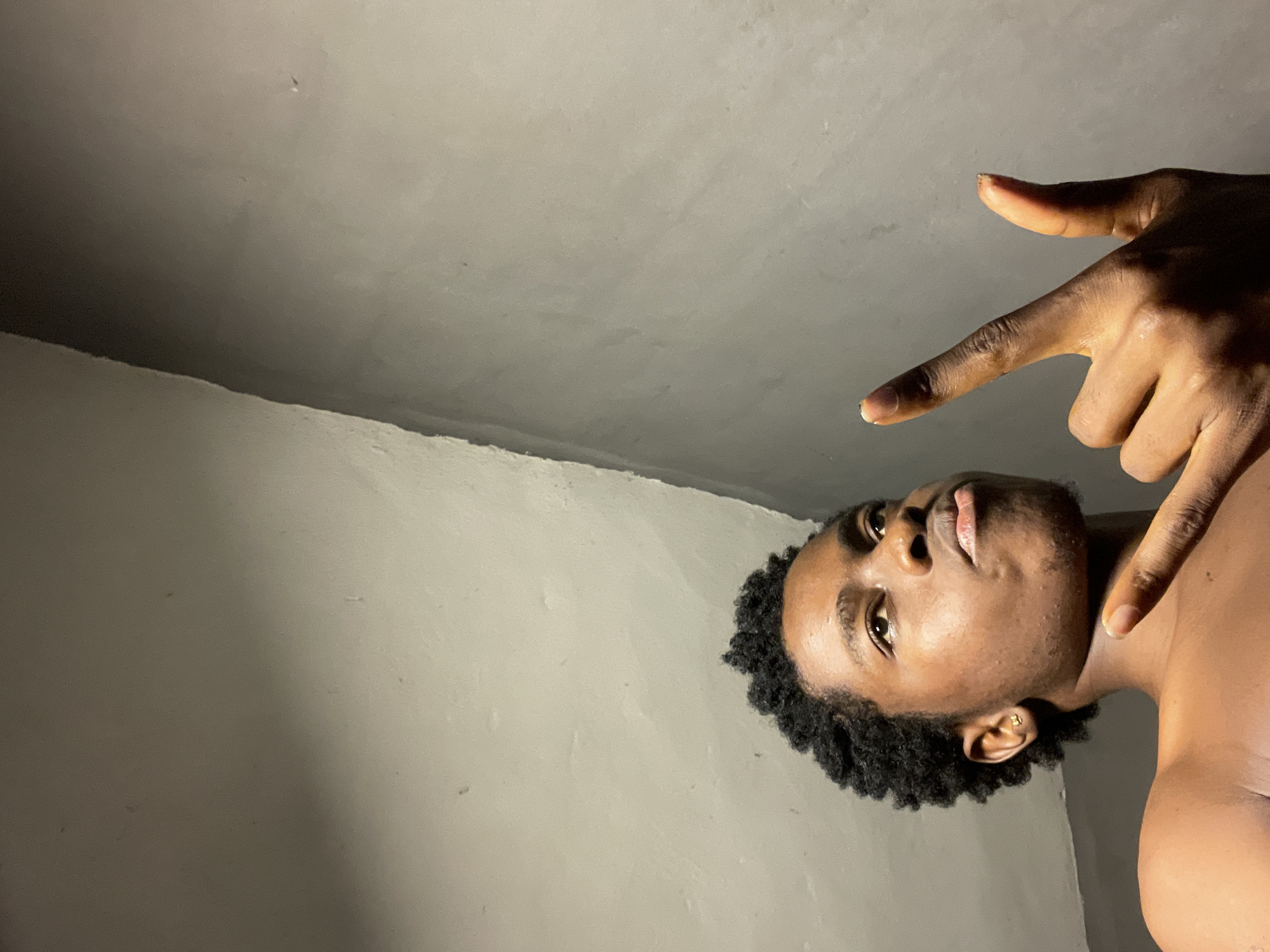 A low-angle photograph shows a shirtless, dark-skinned man with short, curly black hair and a gold earring in his left ear. His face is turned upward, and his gaze is directed toward the camera. His right hand is raised, with the index finger and thumb extended, forming a "finger gun" gesture pointing upwards. The background consists of textured, light gray walls and ceiling. The location in the city of Nkpor, Nigeria is not discernible from the image.