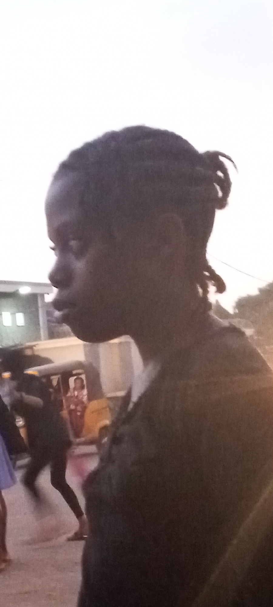 A dark-skinned person with braids is standing in Sabon Tasha, Nigeria. Other people are walking in the background, including a person with bare legs walking away and a person in blue standing to the left. A yellow commercial vehicle, possibly a tuk-tuk, is visible in the background. A building with lighted windows is to the left of the vehicle.