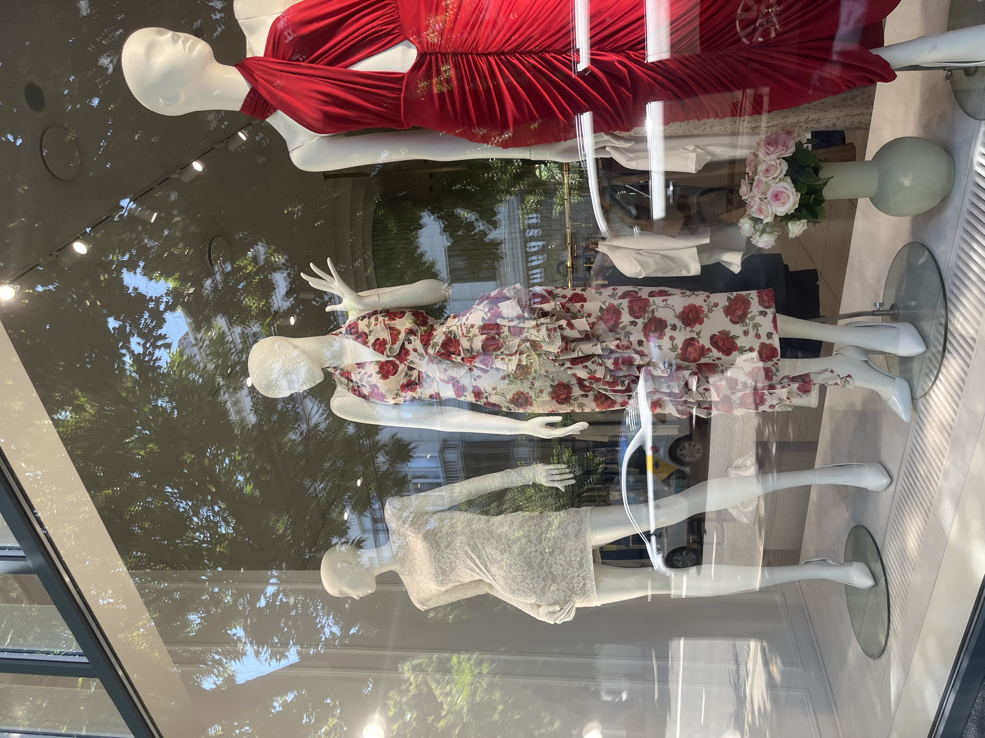 The image shows a window display of a clothing store, likely in Krasnodar, Russia. Three mannequins are displayed, each wearing a different outfit. The top mannequin is wearing a flowing red dress with a ruched bodice. Below it, a mannequin wears a white dress with a floral print featuring red roses and ruffled details. The bottom mannequin is dressed in a short, white lace dress.
The display is set against a backdrop that reflects the outdoors, showing trees and buildings, suggesting it's daytime. The lighting in the display is artificial, with several spotlights illuminating the mannequins. A vase with pink roses is also visible in the display, adding a decorative element. The mannequins are made of white, smooth material, and their poses are static, typical of a retail display. The overall impression is one of a stylish, contemporary fashion presentation. The image is a photograph taken from outside the store, looking through the glass.