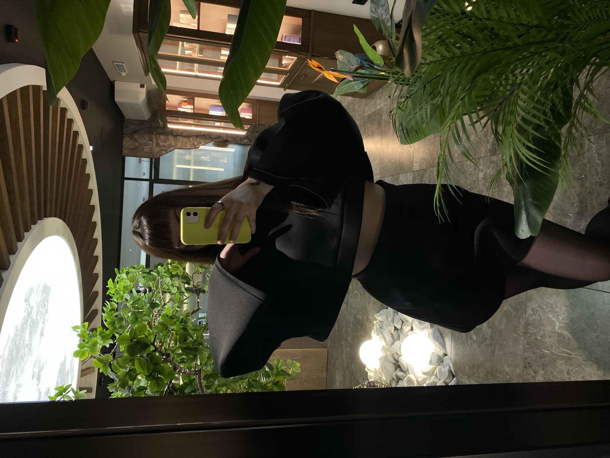 A young adult female is reflected in a mirror. She is holding a yellow cellphone, possibly taking a photograph of herself. She wears a black outfit consisting of a top and a skirt, as well as black tights. The background includes a display case stocked with various items and houseplants. The floor is tiled, and there are decorative stones near a light source. A rounded architectural detail with wooden slats is visible on the left. The location cannot be determined as Votkinsk, Russia, from provided image details alone.