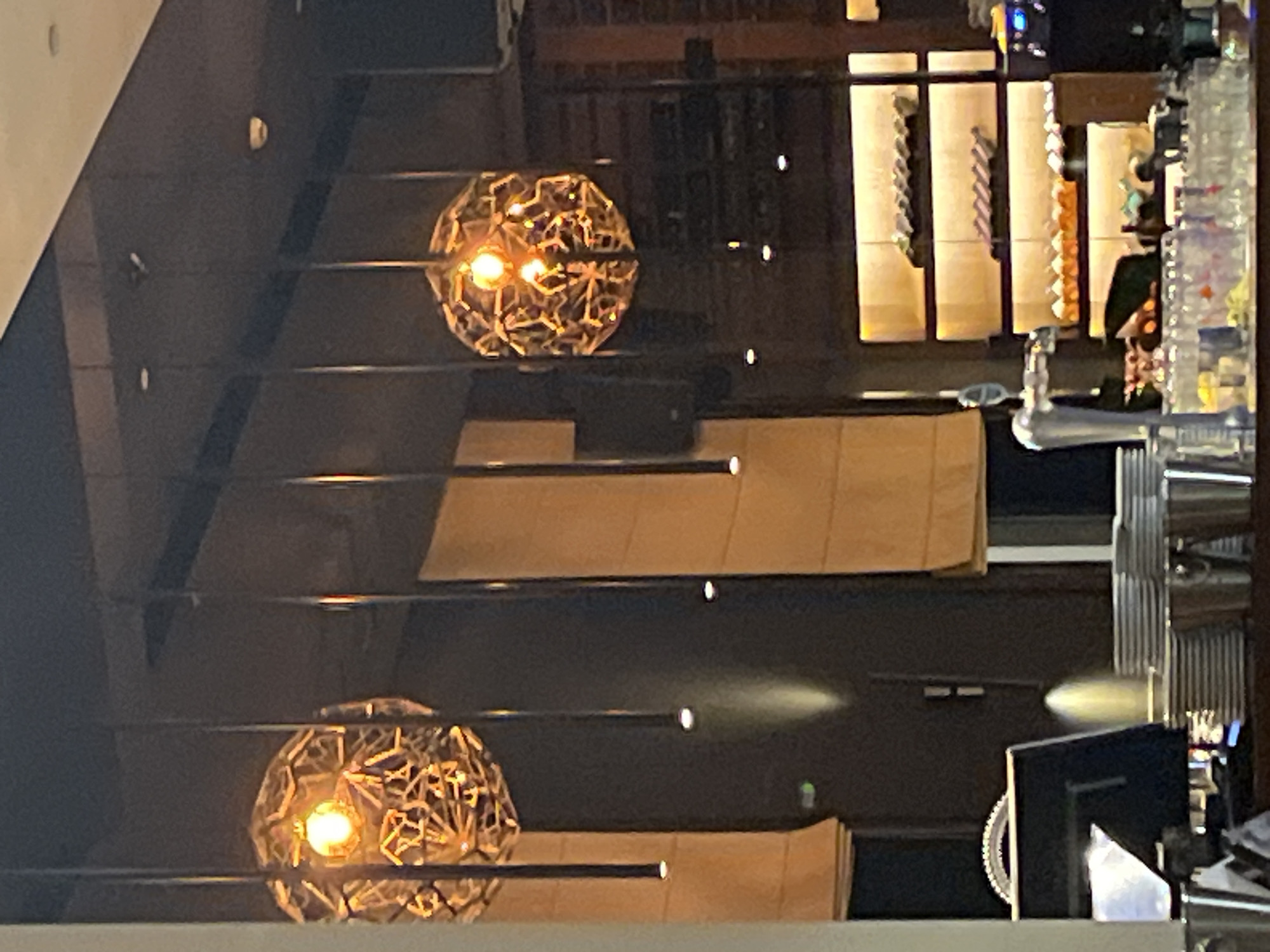 This image depicts the interior of a restaurant or bar. Suspended from the ceiling are decorative, geometric light fixtures with visible bulbs emitting a warm, orange glow. The fixtures are interconnected by thin rods. Below the lighting, a dark wall is partially obscured by beige drapes.
On the left side of the frame, a reflective surface, possibly a counter or table, is visible, with a stack of white plates and a stainless steel bucket or container. Further back, a bar area is partially visible, featuring rows of bottles on shelves illuminated from behind. In the foreground, various glassware and bottles are arranged on a counter. A chrome beer tap is also prominently featured. A person is partially visible on the far left of the image, wearing a white shirt.
The overall ambiance is dimly lit with warm accent lighting, suggesting an evening or indoor setting. The specific location of Budva, Montenegro is not discernible from the visual information provided in the image.