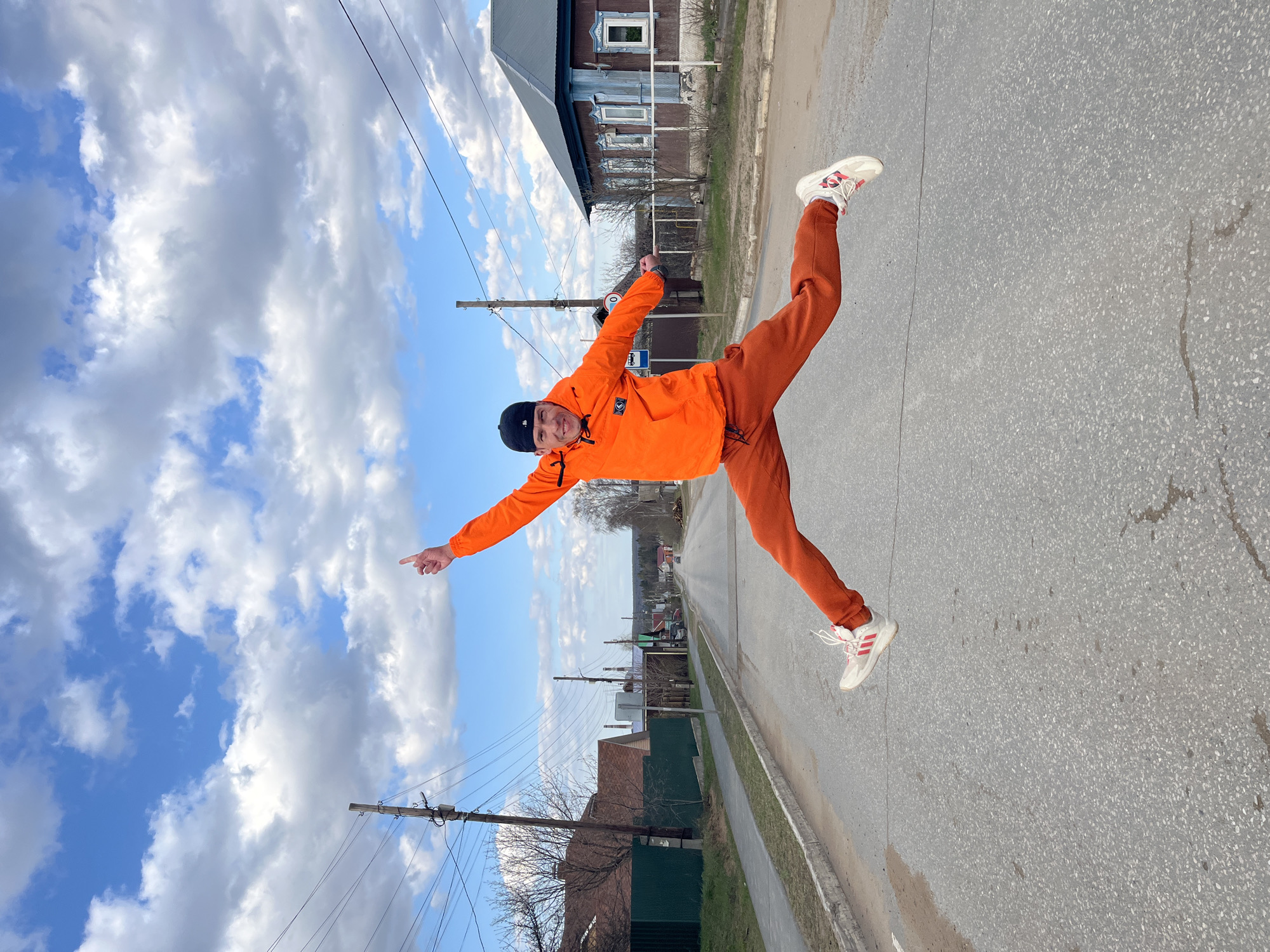 This image captures a young man dynamically posing outdoors on a sunny day in what appears to be a street in Chaikovsky, Russia. The central subject, a man, is depicted mid-action, possibly jumping or balancing on one foot, with his left leg bent upwards and his right leg extended. Both of his arms are stretched horizontally, and his left hand is pointing outwards. He is smiling broadly, conveying a sense of energy and cheerfulness.
He is dressed in a striking bright orange hooded jacket and matching orange track pants, paired with a black baseball cap and white sneakers featuring red accents.
The setting includes an asphalt road in the foreground and midground, with a visible curb. The background features traditional-style wooden houses, some with intricate window details, along the side of the street. Utility poles with multiple wires crisscross the upper portion of the scene. The sky is a vibrant blue, adorned with scattered white and light grey clouds, indicating clear daytime weather. The vibrant orange of the man's outfit stands out against the more subdued tones of the street and buildings. There is no discernible text visible within the image.