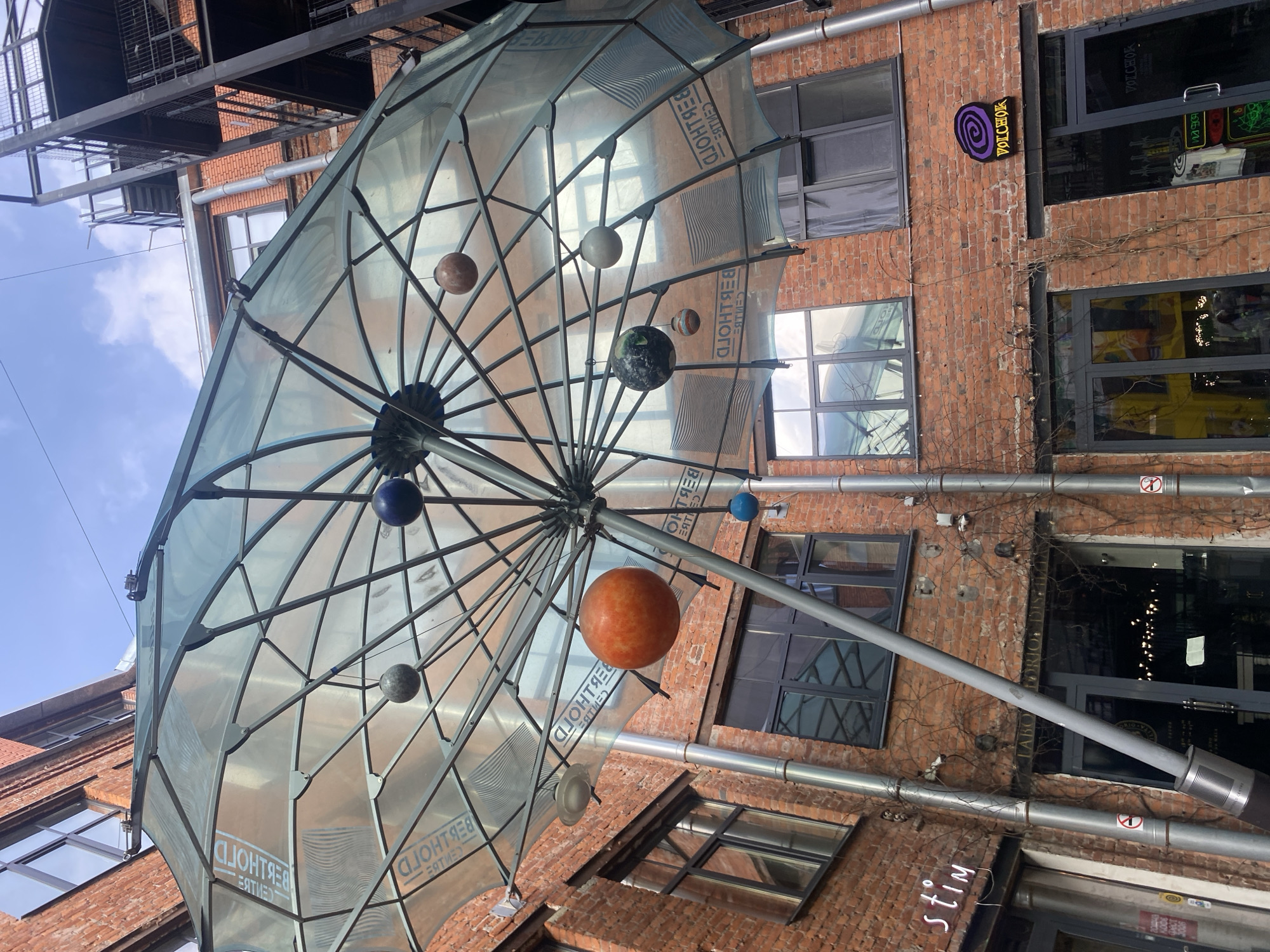 The image captures an architectural scene in Saint Petersburg, Russia. The focus is on a large, decorative umbrella-like structure that appears to be part of an art installation or street decoration. The structure is made of a translucent material stretched over a metal frame and features spherical objects resembling planets suspended inside.
The background consists of a brick building exterior with various windows and pipes running along the walls. Graffiti and signs, including one that reads "VOLCHOK," are visible on the building. There is also a sign reading "stim". The sky is partially visible in the upper left corner of the frame.