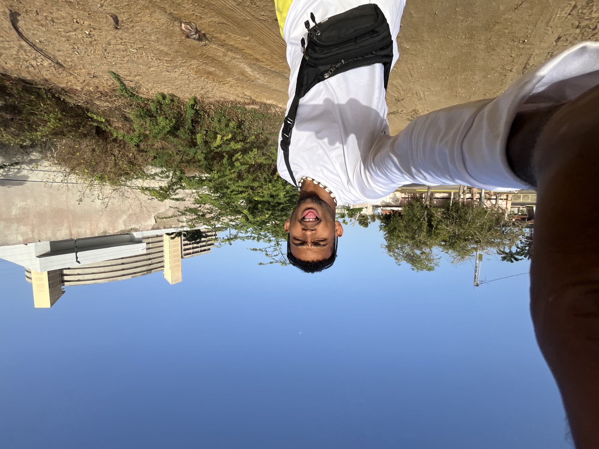 The image is an upside-down selfie of a man with tan skin, short black curly hair, and a beaded necklace. He is wearing a white T-shirt, a black fanny pack across his chest, and yellow shorts. His tongue is slightly sticking out. The background includes a clear blue sky, and a building with a curved design, and some greenery. There is also construction equipment including a crane. The ground is dirt and vegetation. The location is likely Porlamar, Venezuela, based on the environment.