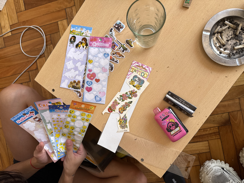 A top-down shot on a wooden floor reveals a light brown tabletop. The table holds sticker sheets featuring dogs, hearts, weapons, temporary tattoos, and smiley faces. A glass, an ashtray filled with cigarette butts, rolling papers, and a pink vape pen also sit on the table. A person holds sticker sheets with Hello Kitty and star designs in their hands. A white cable lies beside the table, and designer sneakers are visible at the table's corner.