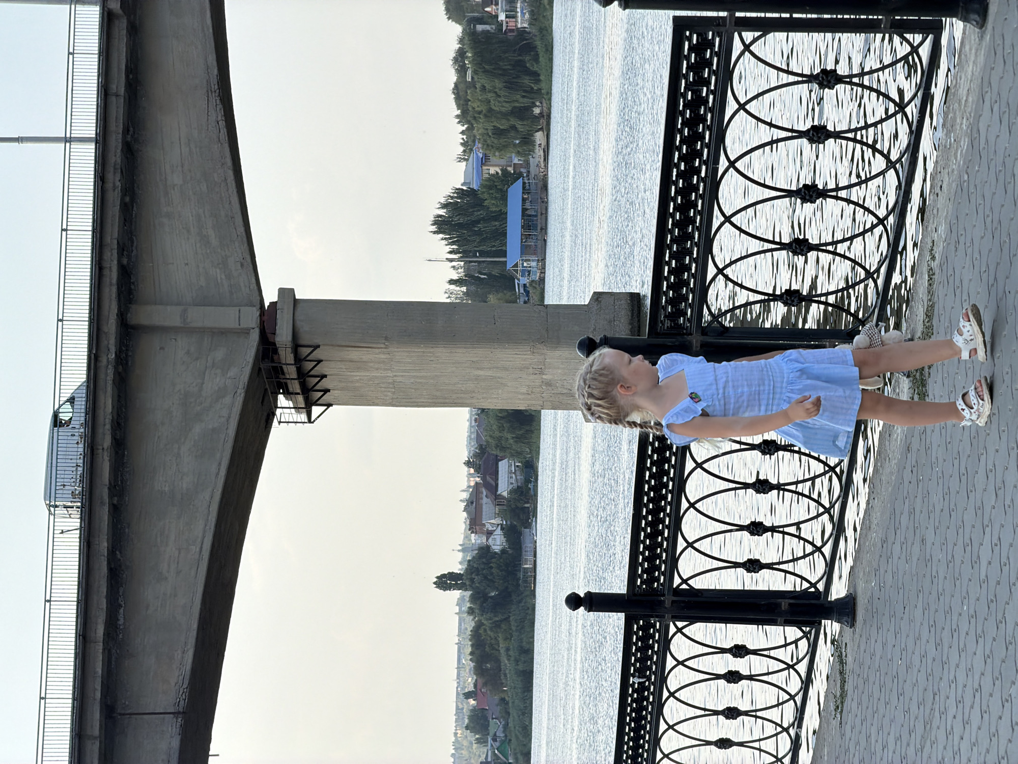 A young blonde girl wearing a blue dress and sandals is walking on a paved pathway next to a body of water, likely the Volga River, in Kamyshin, Russia. She is holding a stuffed animal. A decorative black metal fence separates the pathway from the water. In the background, across the river, is a town with buildings and trees. A concrete bridge, part of the Kamyshin Bridge, looms overhead with a white van visible on it. The sky is overcast.