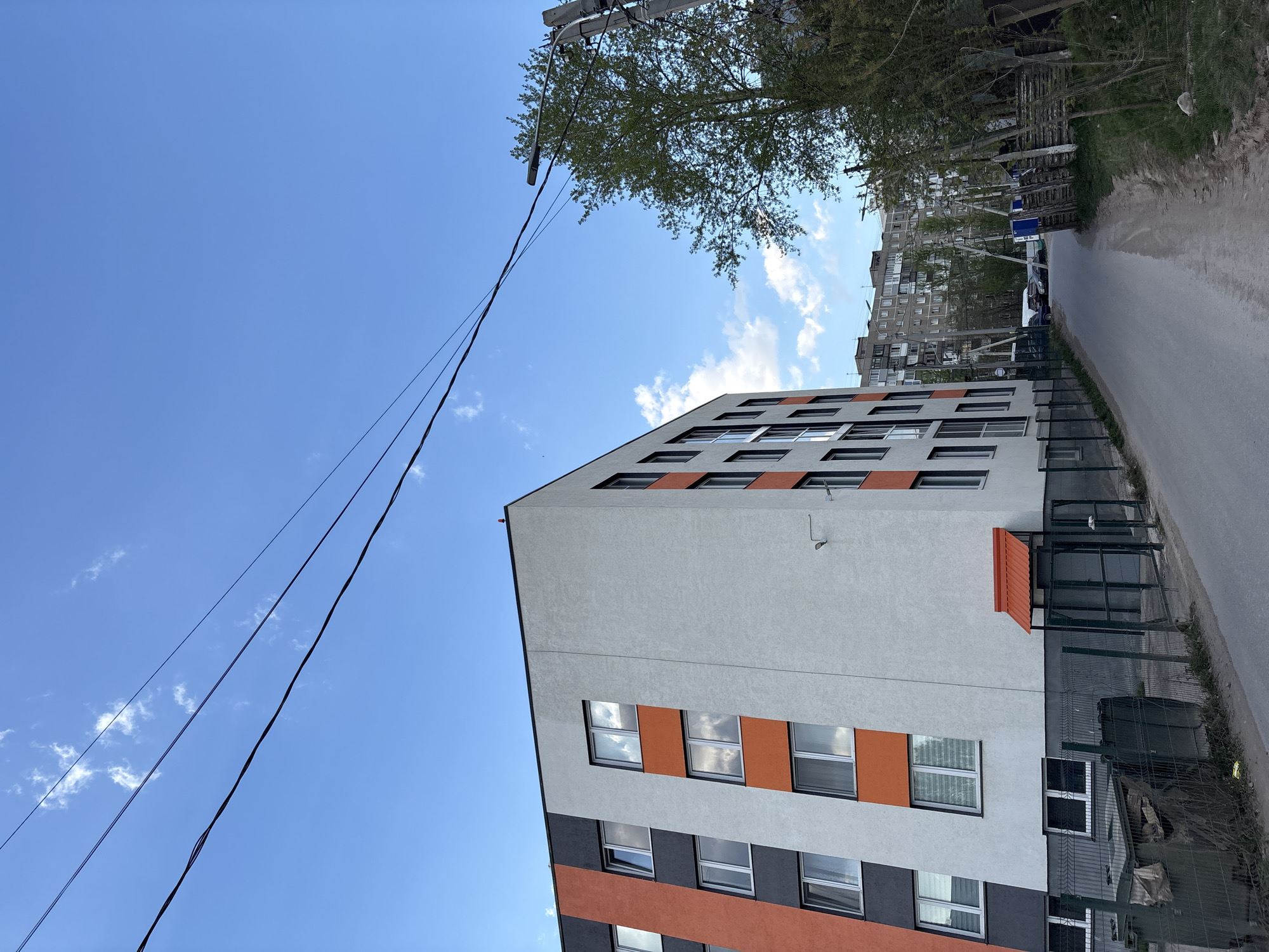 The image captures an upward-angled view of a modern apartment building in Perm, Russia, under a clear blue sky with scattered clouds. The building has a multi-toned facade featuring sections of grey, black, and orange. Windows are arranged in a grid pattern. A small orange awning is visible above a ground-level entrance. Power lines stretch diagonally across the upper-left portion of the frame, connecting to a utility pole obscured by foliage. In the background, another older apartment building can be seen along a narrow, paved road lined with trees and street signs. A green chain-link fence partially obscures the lower-right section of the building. There are no people visible in the scene.
