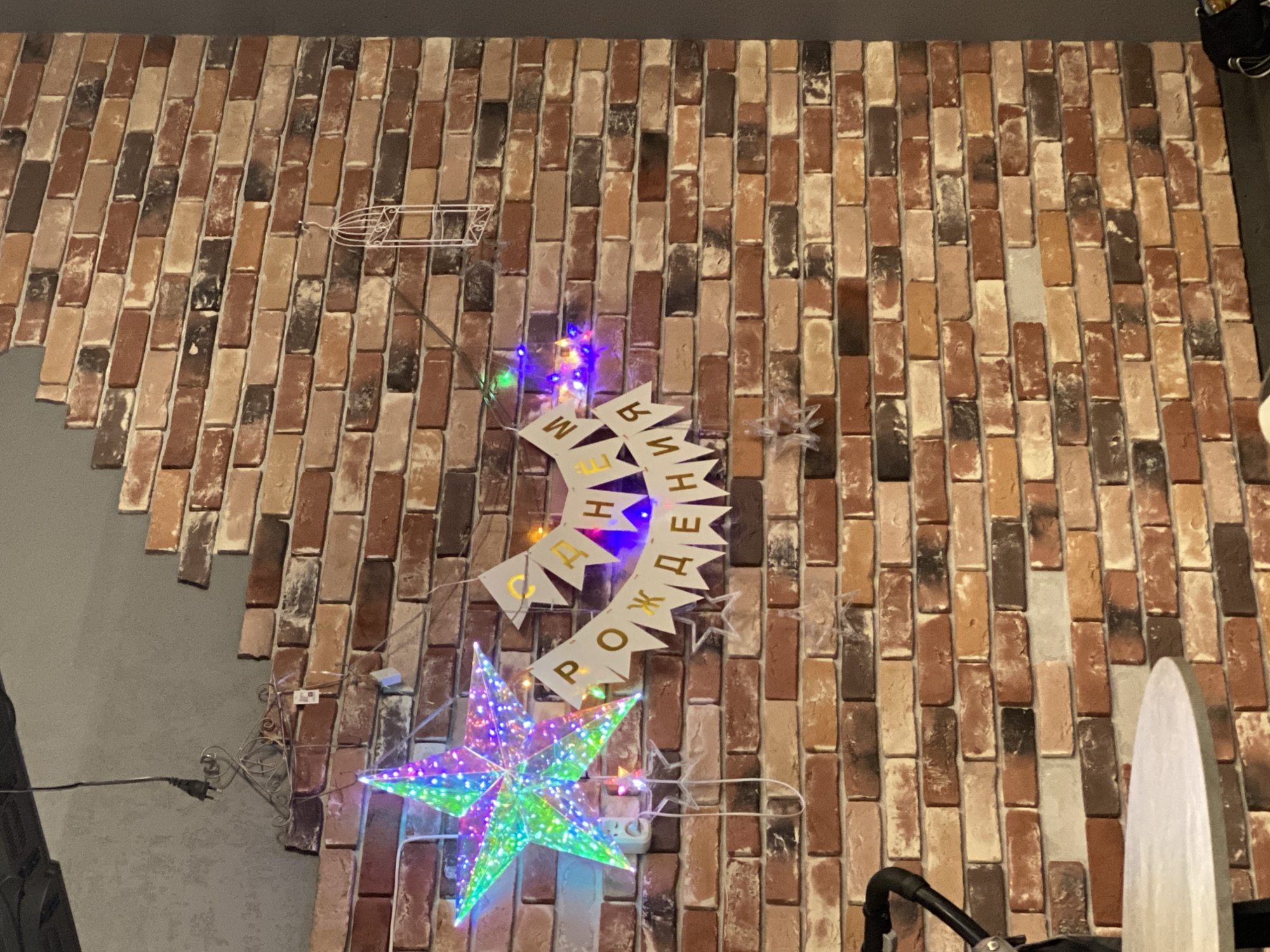 A close-up, eye-level shot of a brick wall in Мирный, Russia, decorated with a festive birthday banner and lights. A large, multi-colored star-shaped light is prominently displayed on the left, with a string of colorful LED lights draped across the banner. The banner itself consists of triangular flags with golden Cyrillic letters spelling out "С ДНЕМ РОЖДЕНИЯ" (Happy Birthday). The brick wall has a rustic, textured appearance, with varying shades of red, brown, and beige bricks. In the upper right corner, a small, white, decorative birdcage hangs from a string. A part of a stroller is visible in the lower left corner, and a round tabletop is partially visible in the bottom left. The overall scene suggests a birthday celebration in a room with a faux brick wall.