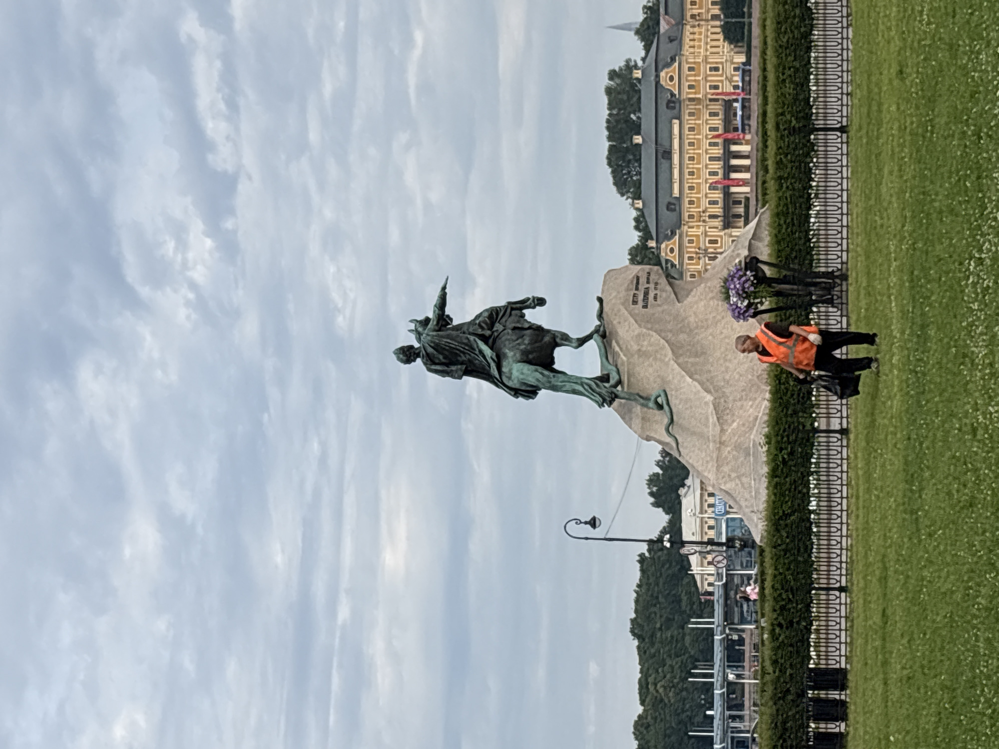 A man in an orange vest walks across a grassy area in front of a large equestrian statue on a stone pedestal, with a building in the background. The statue is made of bronze and depicts a man on a horse, with the horse rearing up. The man on the horse is pointing his right arm forward. The stone pedestal has an inscription on it. In the background, a large, yellow building with a dark roof is visible. The building has many windows and a central entrance. There are trees and a fence in front of the building. The sky is cloudy. The man in the orange vest is wearing black pants and is carrying a black bag. There are flowers in a planter near the statue.
