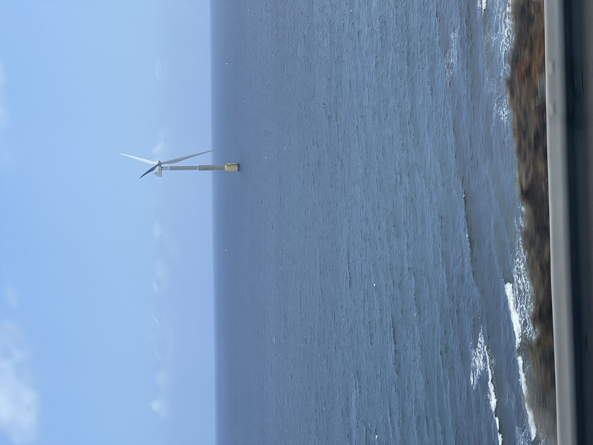 The image, when viewed upright, depicts an offshore wind turbine standing in a vast expanse of the ocean under a clear blue sky, likely off the coast of Telde, Spain.
In the mid-ground, a tall, slender wind turbine dominates the scene. It features three white, pointed blades, with one pointing towards the sky and the other two angled downwards. The turbine's tower is a light gray, supported by a structure that includes a distinctive yellow, rectangular component at its base, suggesting its offshore foundation.
The ocean occupies a significant portion of the frame, stretching from the horizon to the foreground. The water is a deep blue-gray, exhibiting a textured surface of countless small ripples and waves, indicating a dynamic but not stormy sea. To the far right, a sliver of coastline is visible, appearing as a dark, rugged strip of land where white foamy waves gently break against the shore.
The sky, a bright and uniform light blue, fills the upper section of the image, with only subtle, diffuse white clouds barely perceptible. The overall setting is a clear daytime scene with good visibility. No people or visible text are present. The scene highlights the integration of renewable energy infrastructure with the natural marine environment.
