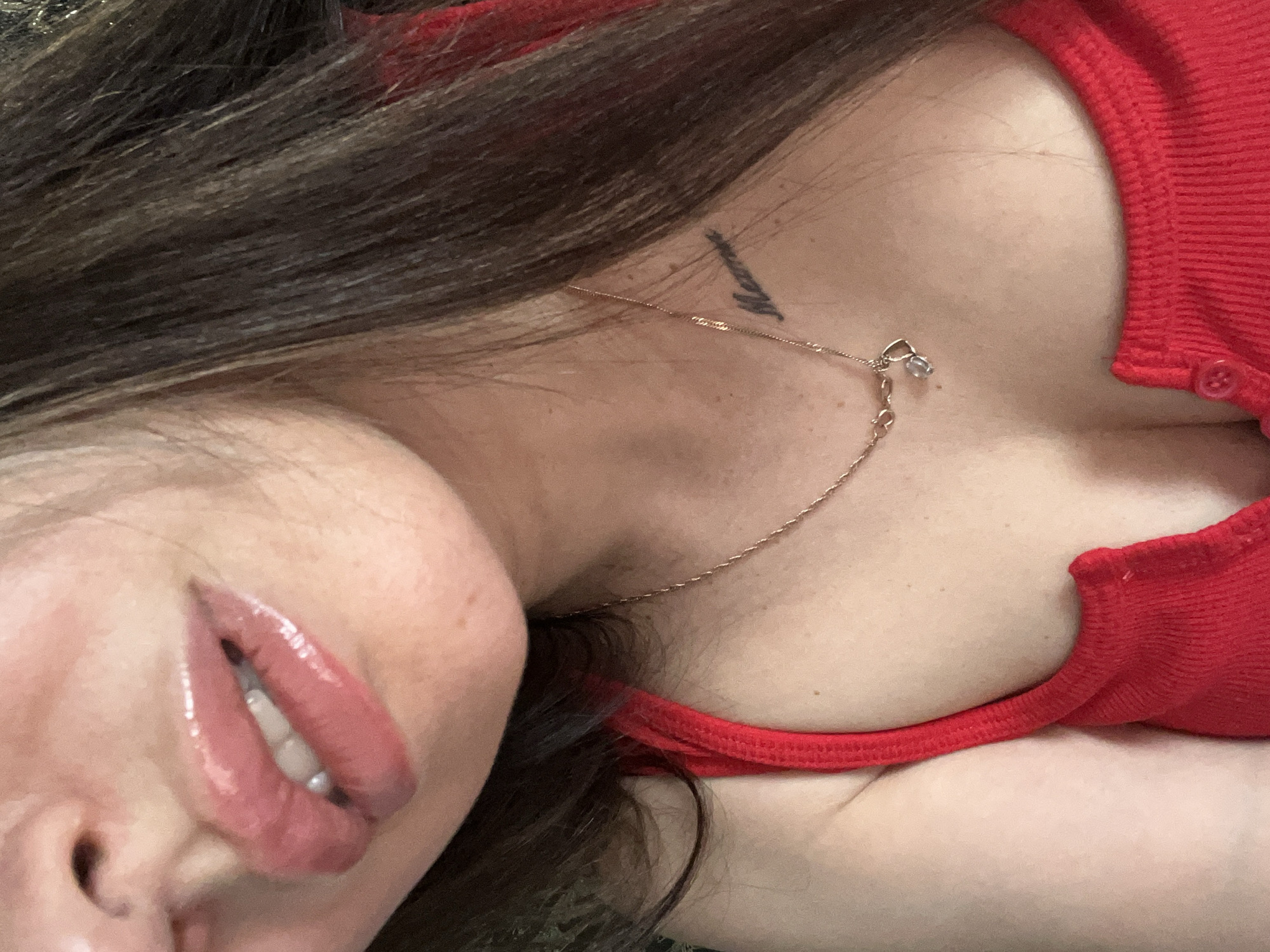 A close-up, mid-shot photograph captures the lower face, neck, and upper chest of a female subject, rendered in a warm color palette. The subject's full lips, coated in a high-gloss, light pink product, are slightly parted, revealing a portion of her white upper teeth. Her skin tone is fair, exhibiting minor texture and faint freckles on the chest. Long, dark brown hair frames the right side of her face and cascades over her right shoulder and upper chest, partially obscuring a small, dark script tattoo on her right collarbone area that appears to read "Mama." She is wearing a red, ribbed tank top or camisole with a visible button closure near the center of the chest. A delicate gold-colored chain necklace adorns her neck, featuring two small pendants: one resembling a heart and the other a clear or light-colored faceted stone. The background is out of focus and indistinct, providing no environmental details. Location: Unknown.