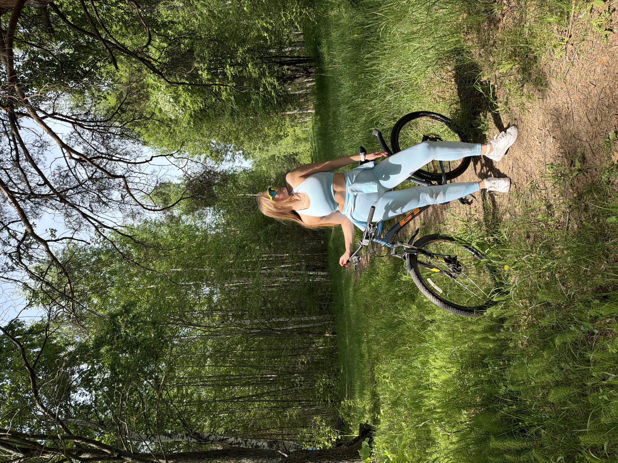 A woman lies diagonally across a bicycle on a dirt path bordered by green grass and trees. The woman is wearing light blue athletic wear and white sneakers, and she is resting on the bicycle's frame. The bicycle is a mountain bike with black tires and a blue frame. The path is surrounded by a forest with tall trees and green foliage. The lighting suggests it is a sunny day. The scene is likely located in a park or wooded area in or near Chaikovsky, Russia, as specified.