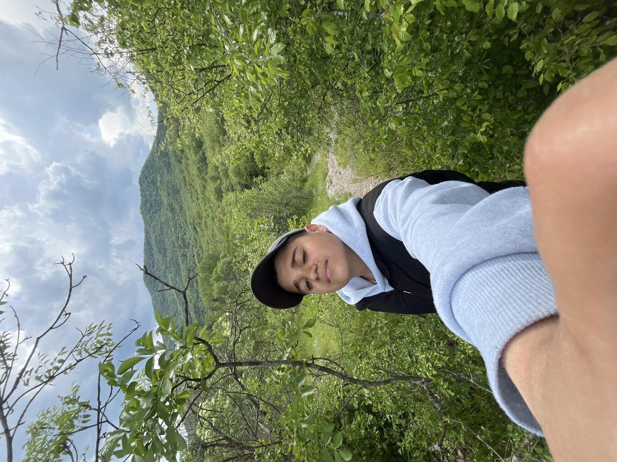 A photograph shows a young individual in a natural setting. The person is wearing a cap, a light-colored hooded sweatshirt, and a dark vest or jacket. They are positioned in front of dense foliage, including trees and bushes. In the background, a mountain or hillside is visible under a cloudy sky. A small pathway can be seen amongst the greenery behind the person. The scene is likely outdoors in a natural environment. The photograph doesn't indicate specific details that could pinpoint a location within Kamchia, Bulgaria.
