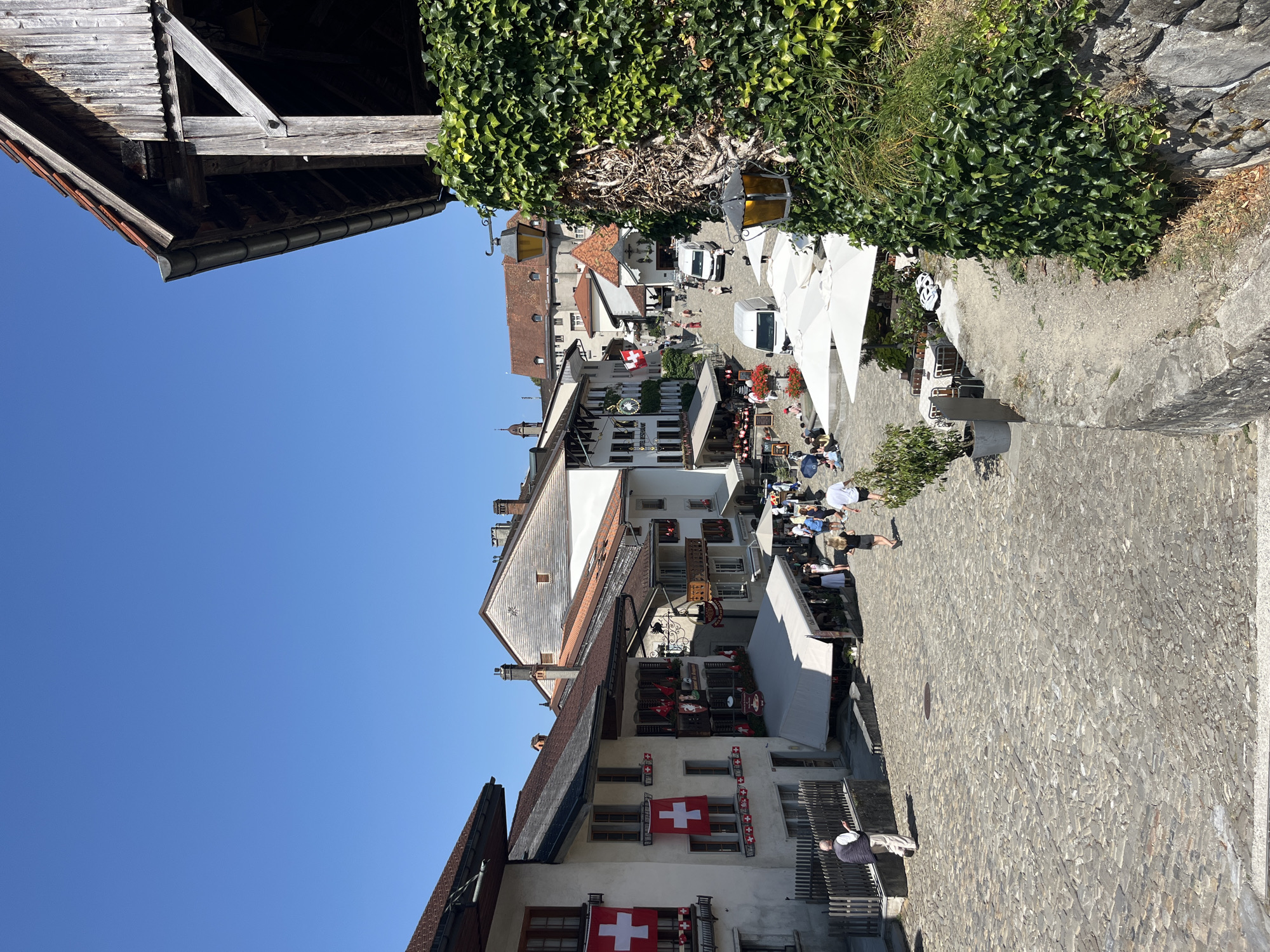 The image depicts a sun-drenched cobblestone square in the town of Epagny, Switzerland, on a clear, bright day. The architecture features traditional Swiss buildings with red-tiled roofs and white facades adorned with numerous Swiss flags. Several buildings house restaurants and cafes with outdoor seating under white umbrellas, indicating a lively atmosphere with patrons enjoying meals. A few vehicles, including white vans, are visible in the square, suggesting local transportation or service. People are seen walking, sitting, and interacting, contributing to the scene's sense of activity. Lush green ivy climbs a stone wall in the foreground, framing the view of the town. The overall impression is a picturesque and bustling European town square during the daytime.