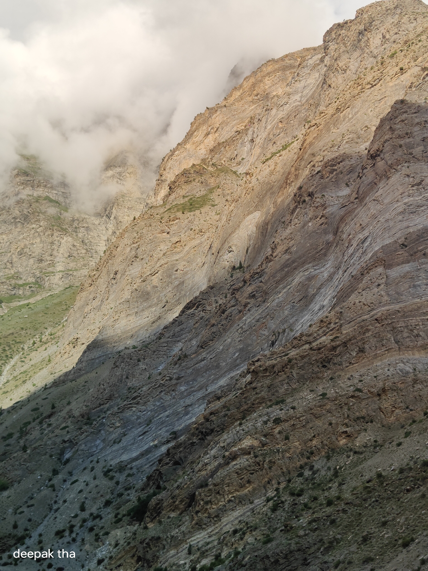 This is a photograph of a mountainous landscape in the vicinity of Gondhla, India. The image captures a steep, rocky mountainside with distinct geological layers. The upper portion of the mountain is bathed in sunlight, revealing shades of tan and yellow rock, with patches of green vegetation on a lower, less steep slope. Wispy white clouds partially obscure the higher peaks, creating a dramatic contrast with the illuminated rock face. A deep shadow stretches across the lower half of the mountainside, highlighting the rugged texture and varied strata. Scattered across the slopes are small, dark green shrubs. The text "deepak tha" is visible in white lettering at the bottom left corner of the image, indicating a potential photographer or owner's mark. The scene appears to be devoid of any human activity or constructed elements.
