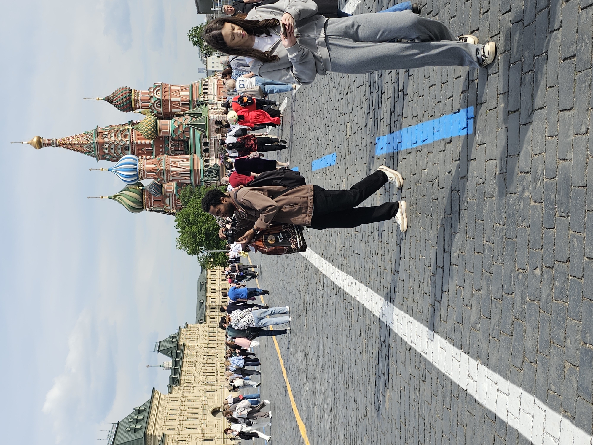 The image shows a wide view of Red Square in Moscow, Russia, with St. Basil's Cathedral visible in the background. Numerous people are walking around the square. A man in a brown jacket and black pants is walking in the middle of the frame, while a woman in a gray tracksuit is standing and looking at her phone. The square is paved with cobblestones marked with a white line and short blue lines. GUM department store is visible on the right. The sky is partly cloudy.