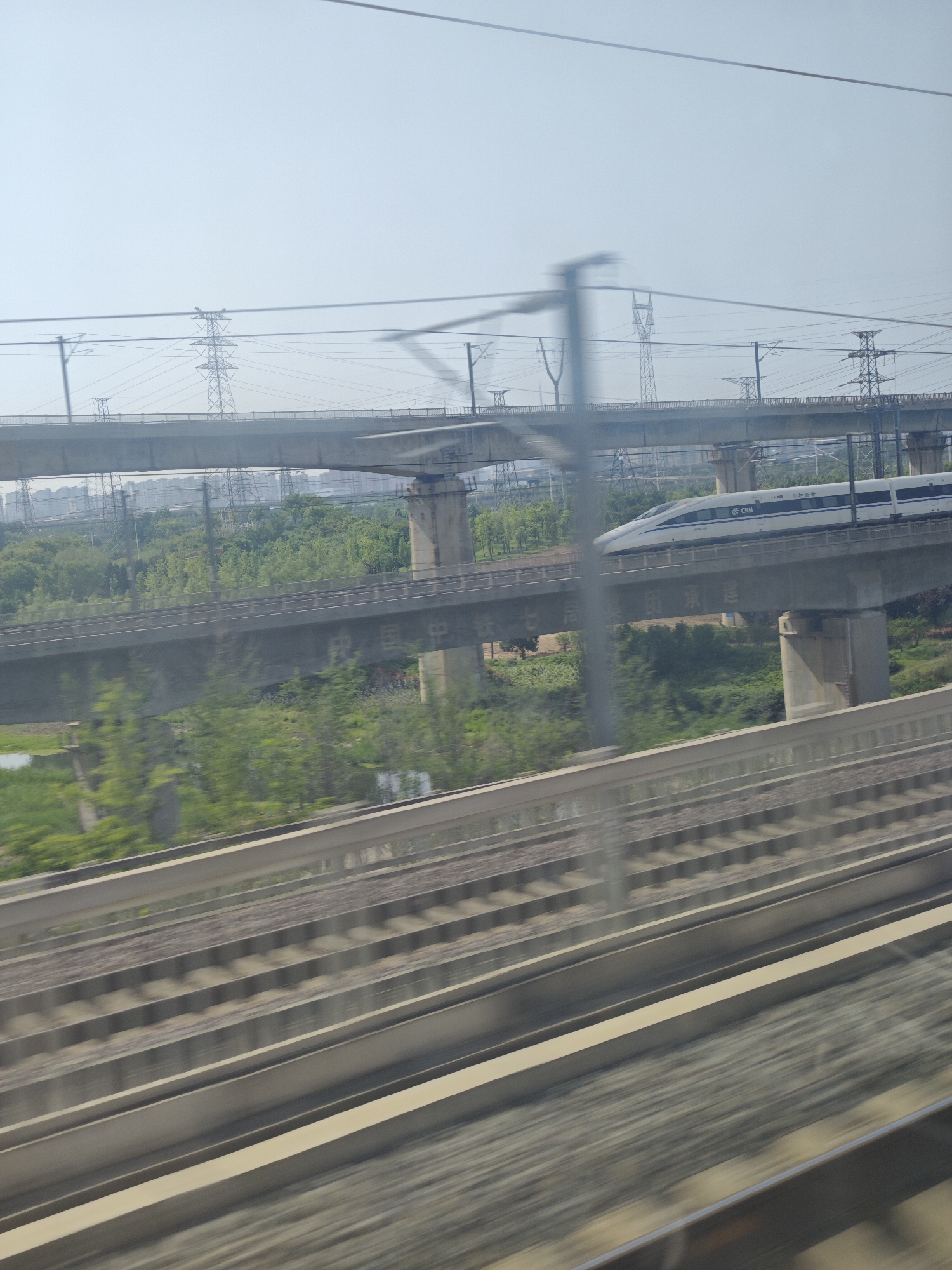 A moving shot from a train window captures the scenery of Unknown, Unknown. The foreground features multiple sets of parallel train tracks, blurred due to the motion of the train. Beyond the tracks, a bridge is visible, with a high-speed CRH train traveling across it. Further in the distance, another bridge runs parallel to the first, with power lines and electricity pylons extending into the sky. A backdrop of greenery and indistinct urban buildings is present. The sky is overcast, casting a soft light over the scene.