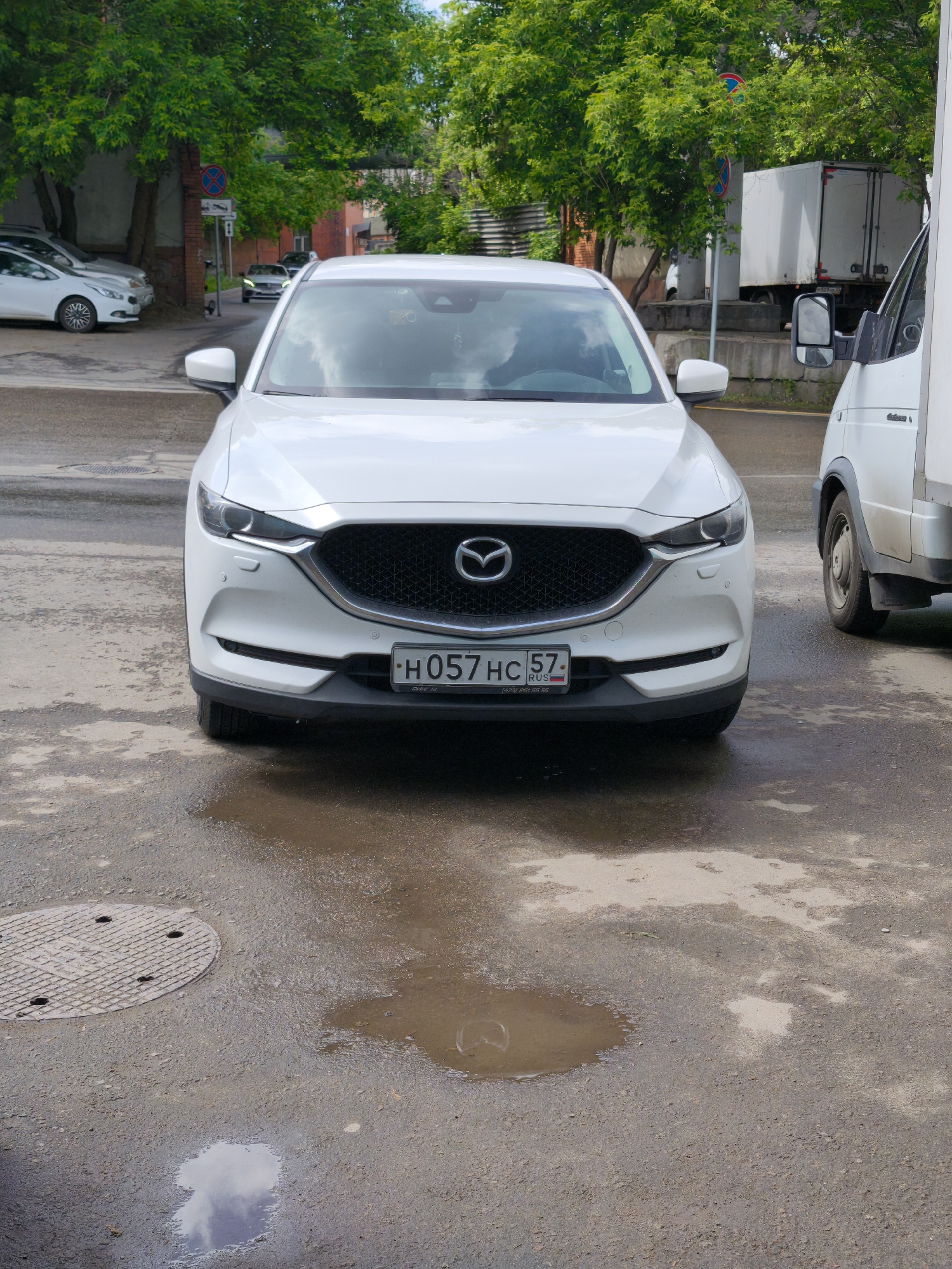 The image shows a white Mazda CX-5 parked on a wet street in Moscow, Russia. The license plate reads "H057 HC 57 RUS". To the right of the Mazda is a white van. Behind the Mazda are several other parked cars. The street is wet and has puddles. A manhole cover is visible on the left side of the road. Green trees and building are in the background.
