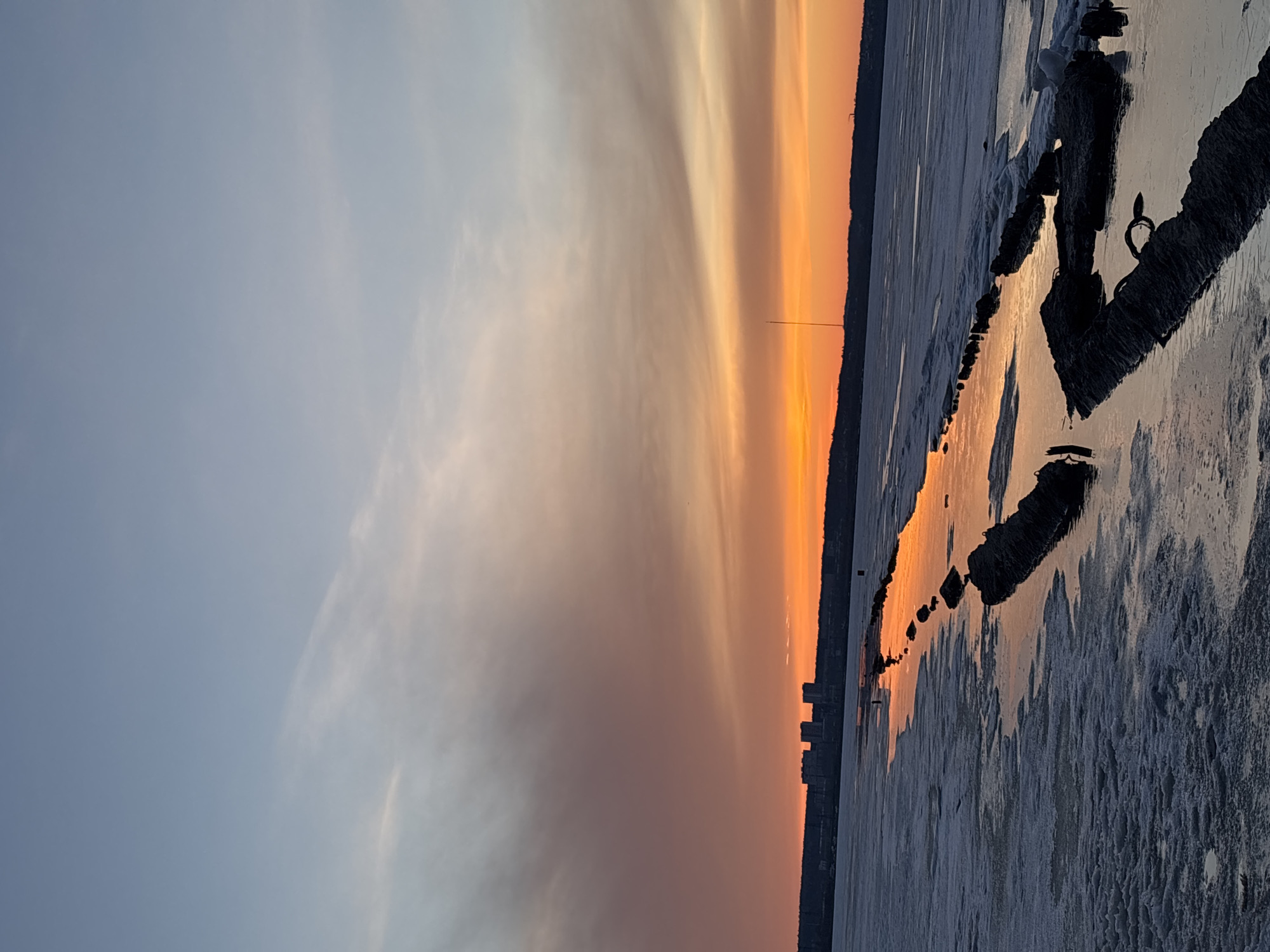 The image depicts a sunset over a frozen body of water, likely a section of the Izhevsk Pond in Izhevsk, Russia, during winter. The sky transitions from a light blue to orange and red hues near the horizon, where a silhouette of buildings is visible. The frozen surface of the pond shows patches of ice and snow, with a dark line of debris or partially submerged objects stretching along the water. The reflection of the sunset light is visible on the icy surface. The overall scene conveys a cold, wintry setting illuminated by the sunset.