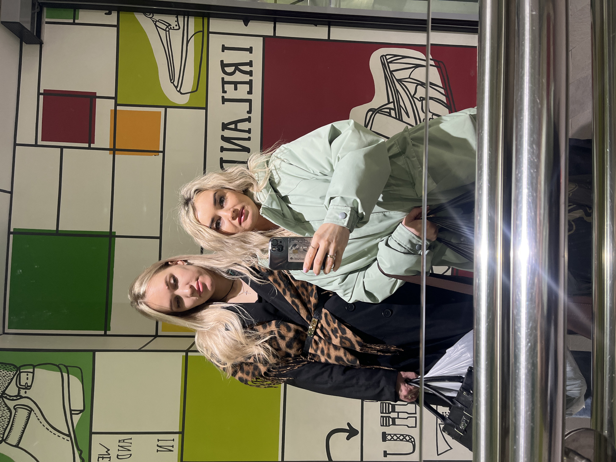 Two blonde women are reflected in a mirror next to a wall in Izhevsk, Russia. The wall has a Mondrian-style design with blocks of green, red, orange, and white, and black lines. The word "BELAVIA" is written backwards on the wall. One woman wears a light green jacket and holds a cell phone in her hand. The second woman wears a black coat with a leopard print scarf. A black purse is visible near the bottom of the reflection, and there is a metal column in the foreground.