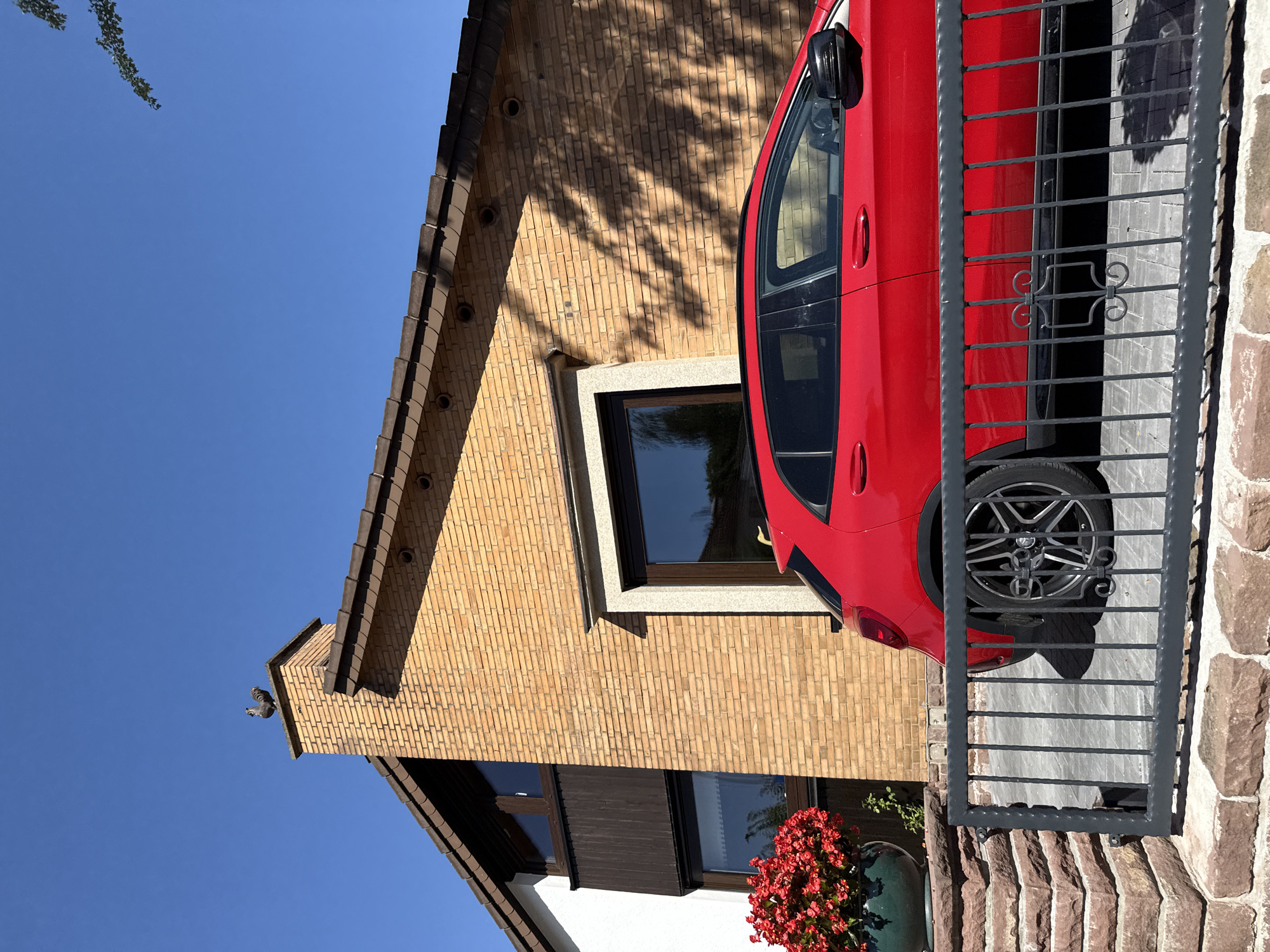 A red car is parked in front of a two-story house with a brick exterior. A rooster figurine is perched on top of the chimney. Red flowers are in a planter to the left of the house. The scene is bathed in sunlight. The location is Bad Sachsa, Germany.