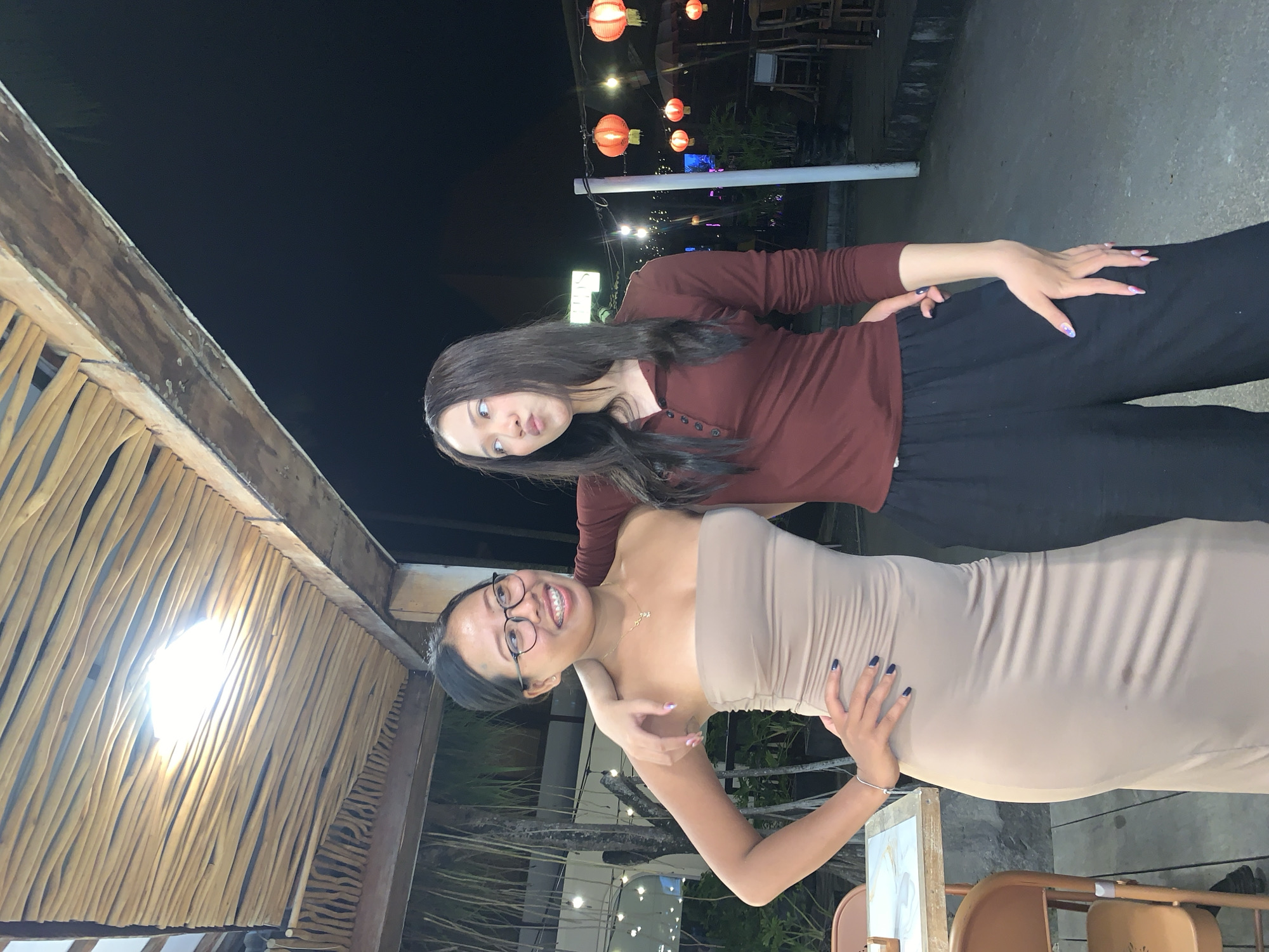 Two young women are standing together outdoors at night. The woman on the left is wearing a tan strapless dress and glasses, and has her arm around the other woman's shoulders. The woman on the right is wearing a maroon long-sleeved shirt and black pants. They are both smiling. The background includes a thatched roof structure, string lights, and red lanterns. The location is likely in North Kuta, Indonesia, based on the architectural details and lighting.