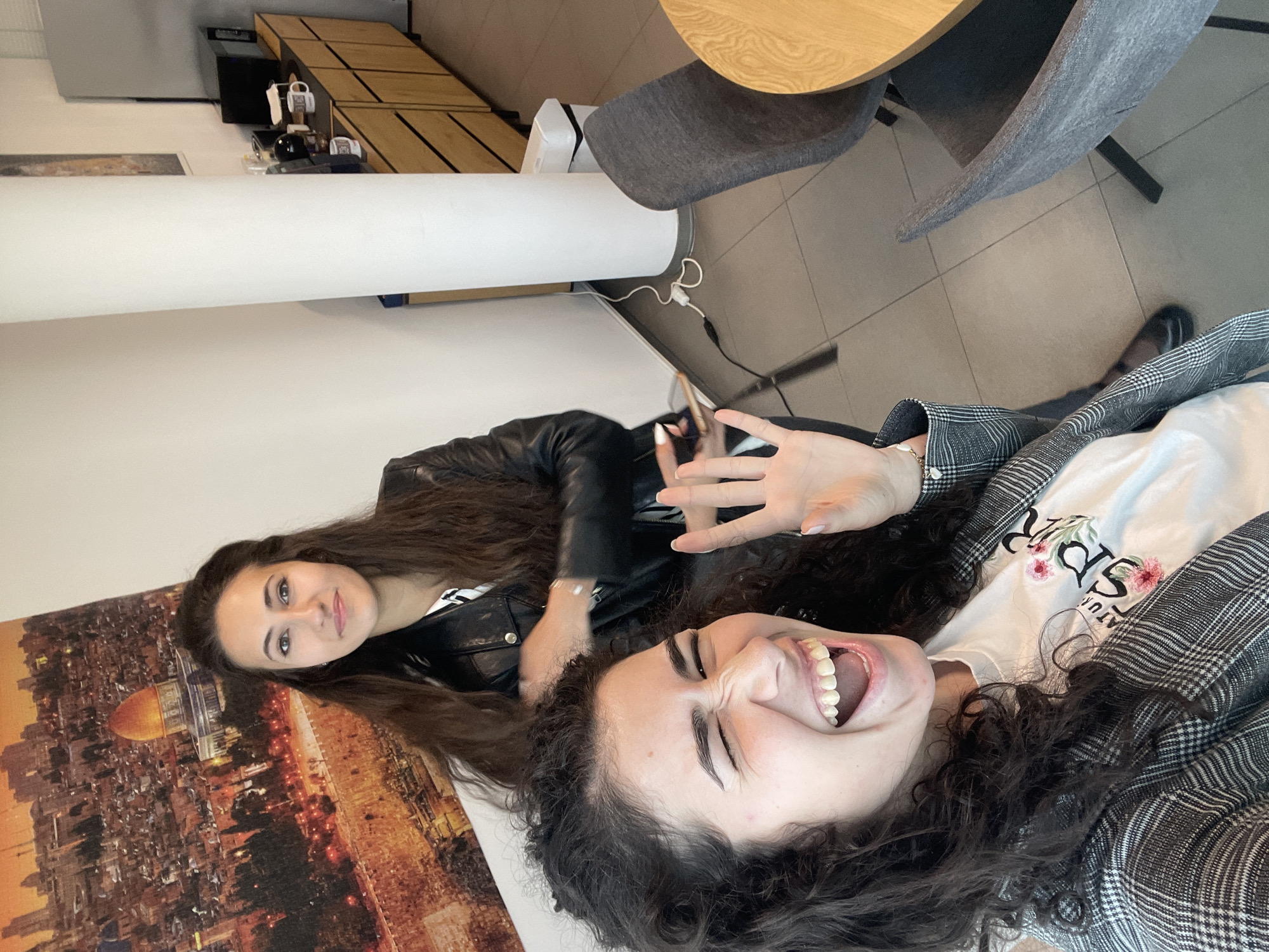 Two young women are lying down and looking at the camera, with one positioned closer to the viewer and laughing while the other is in the background. The woman in the foreground is wearing a white t-shirt with floral designs and a grey plaid jacket. The other woman is wearing a black leather jacket. There is a photo on the wall of the Dome of the Rock in Jerusalem. The women appear to be in a cafe-like setting with a light-colored table and grey chairs. The location is the city of Sofia, Bulgaria.