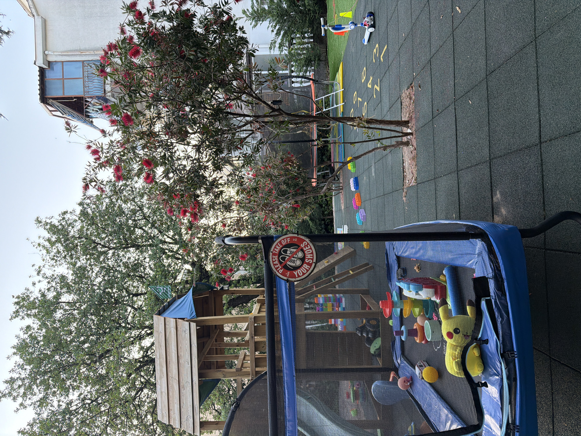 This image captures a vibrant and well-equipped outdoor children's play area, likely in Budva, Montenegro, under a clear daytime sky with soft natural light, possibly morning or late afternoon. The scene features a dark grey interlocking rubber tiled ground, characteristic of playgrounds, adorned with yellow painted numbers like "0," "1," "2," "3," "4," and "5," suggesting games like hopscotch.
In the foreground, a prominent blue trampoline with a safety net is seen. Inside the net, various toys are scattered, including a yellow Pikachu plushie and other colorful plastic items such as building blocks, rings, and a plane-like toy. Attached to the trampoline's safety net is a round, red and white sign that explicitly states, "PLEASE TAKE OFF YOUR SHOES," accompanied by an icon of crossed-out shoes.
To the left, a wooden play structure or treehouse with stairs and platforms is partially visible, blending into the surrounding foliage. The area is rich with greenery, featuring a distinctive bottlebrush tree (Callistemon) with bright red, brush-like flowers, and a large deciduous tree with green leaves providing shade. In the background, a white multi-story building with windows and a balcony is visible, suggesting a residential or resort setting. On the rubber tiles near the trampoline, a blue and white child's scooter (featuring Paw Patrol characters) and colorful building blocks are placed. The scene is devoid of people, presenting a peaceful environment awaiting activity.